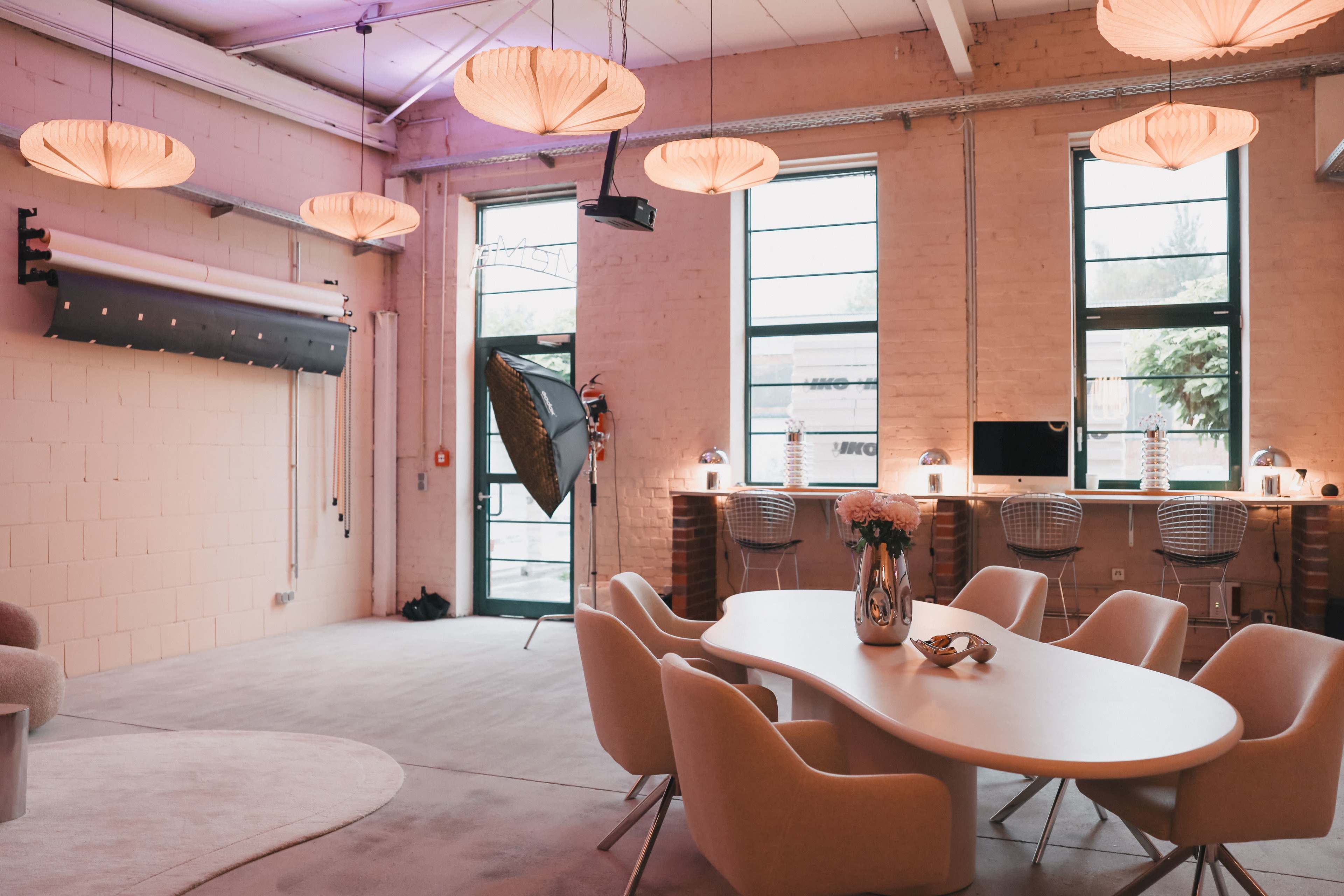 A spacious studio features a circular table surrounded by chairs, with a lighting setup and large windows allowing natural light in.