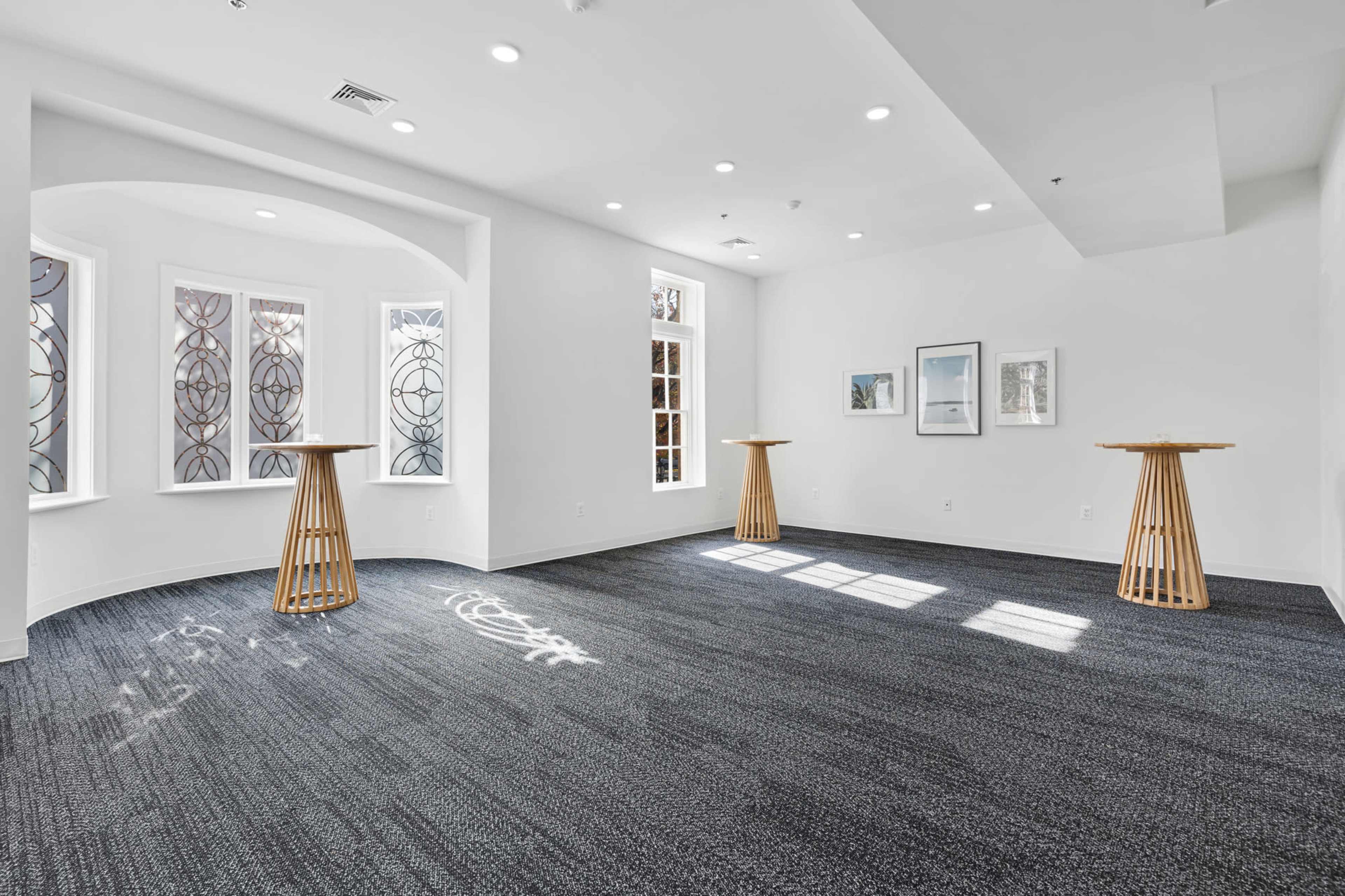 The image shows a bright, empty room with three wooden tables and windows featuring decorative glass patterns.