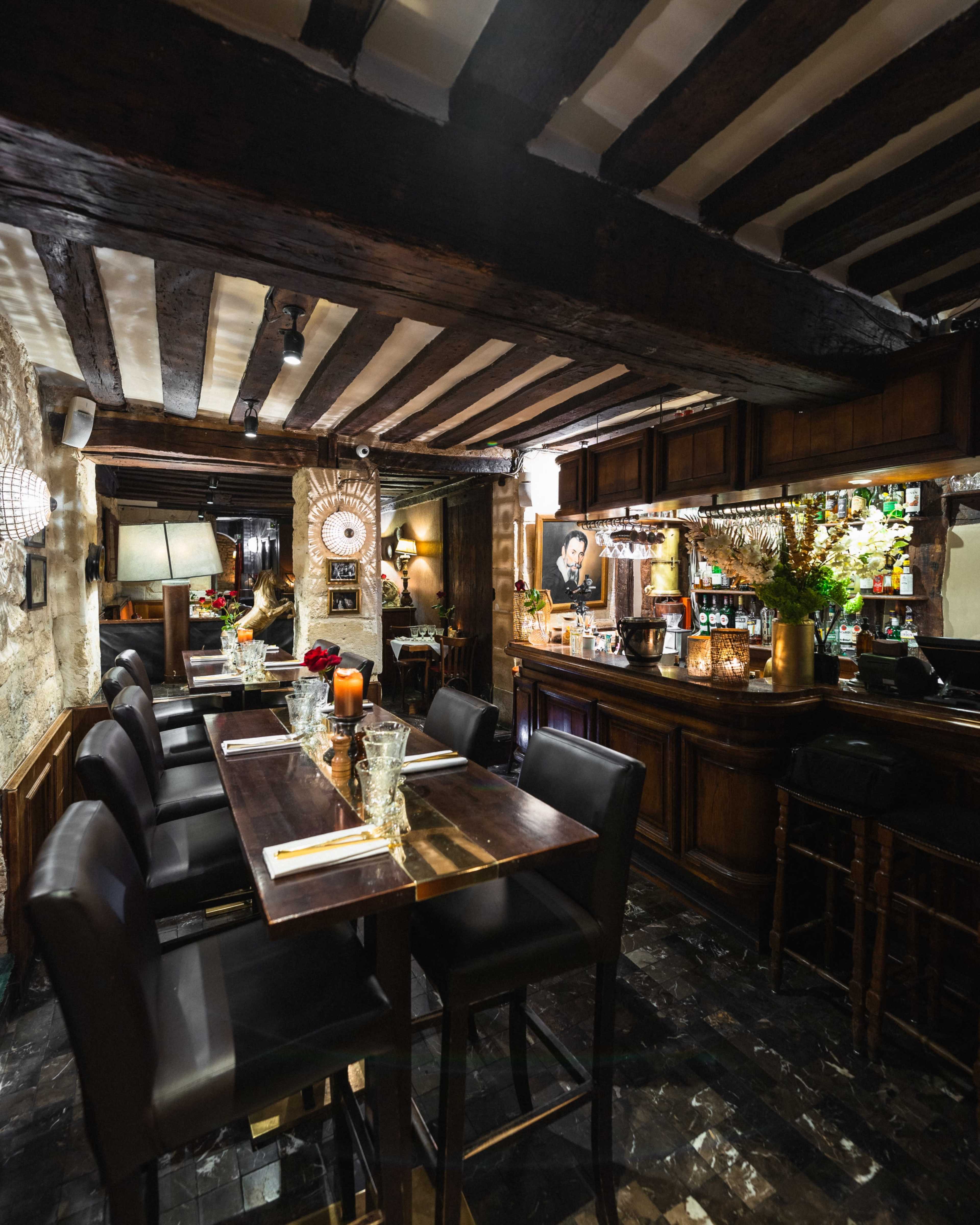 The image shows a dimly lit restaurant interior featuring wooden beams, a bar with various bottles, and tables set for dining.