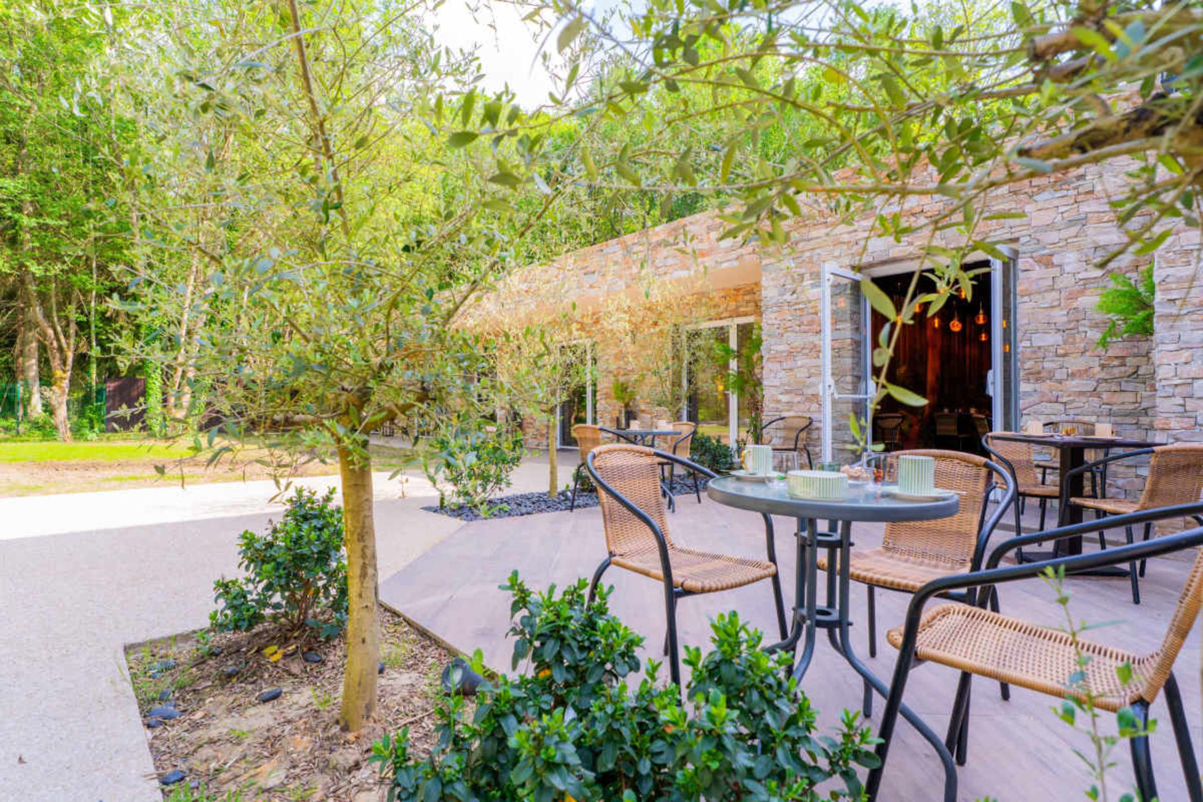 A stone building with large windows is surrounded by greenery and outdoor seating arrangements.
