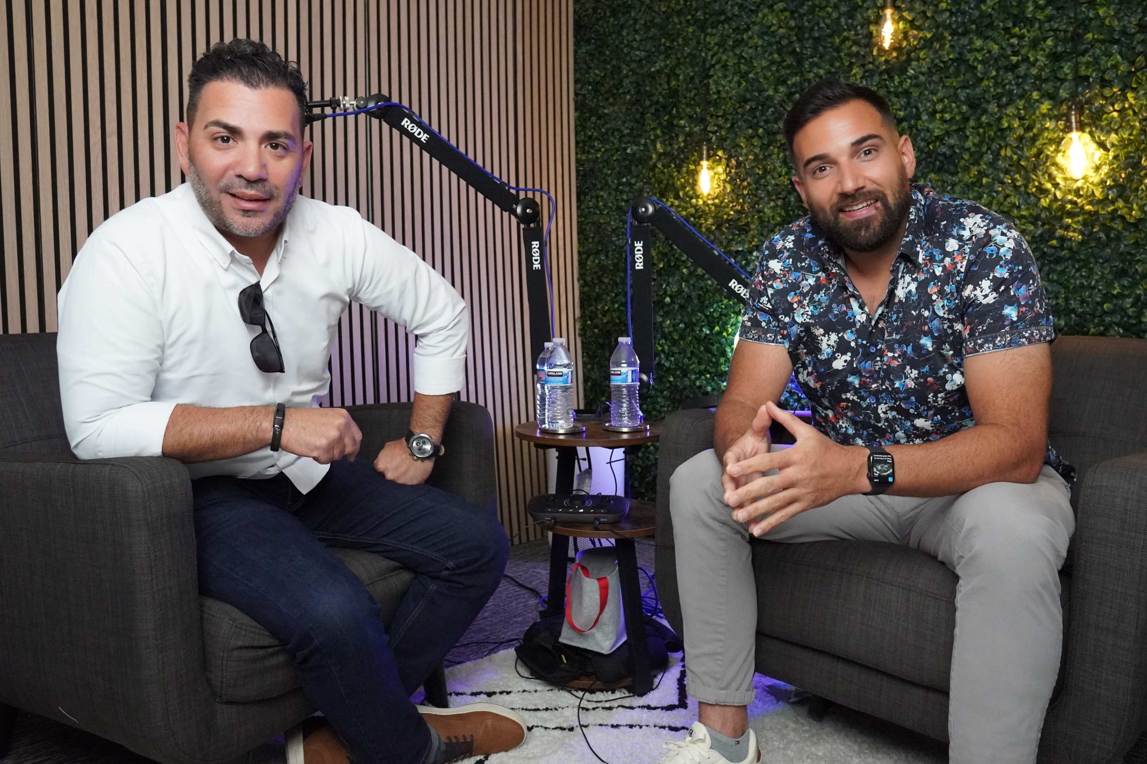 Two men sit in upholstered chairs in a cozy studio setting, with microphones positioned in front of them and a green wall behind.
