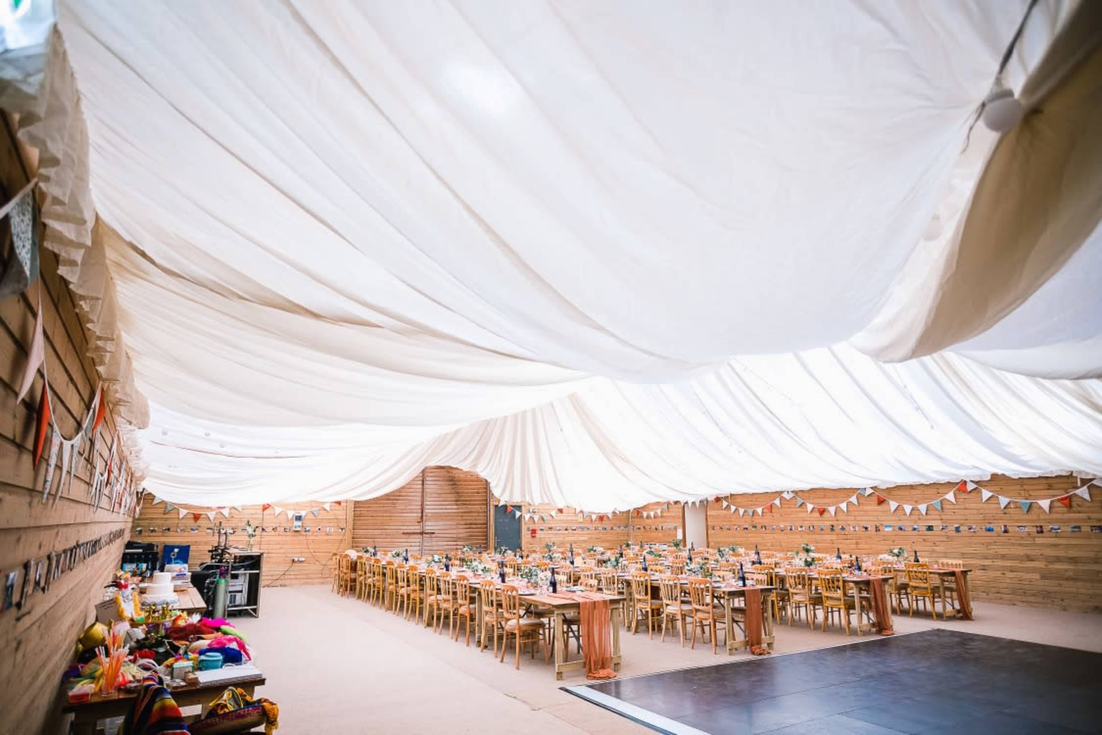 A spacious event space is decorated with a draped white ceiling and arranged with long wooden tables set for dining.