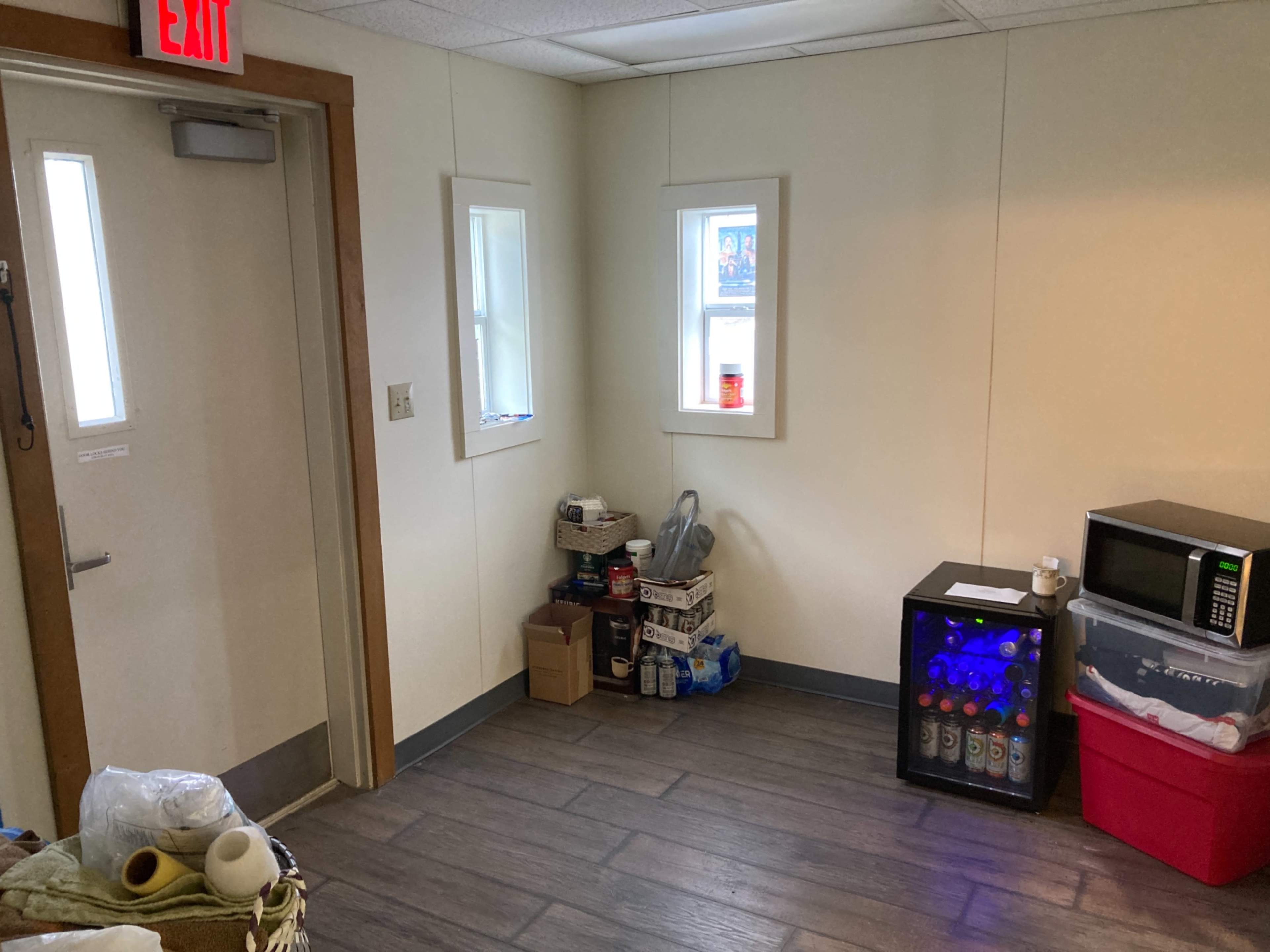 A small corner of a room with a door, two windows, a microwave, a small beverage cooler, and various boxes stored on the floor.