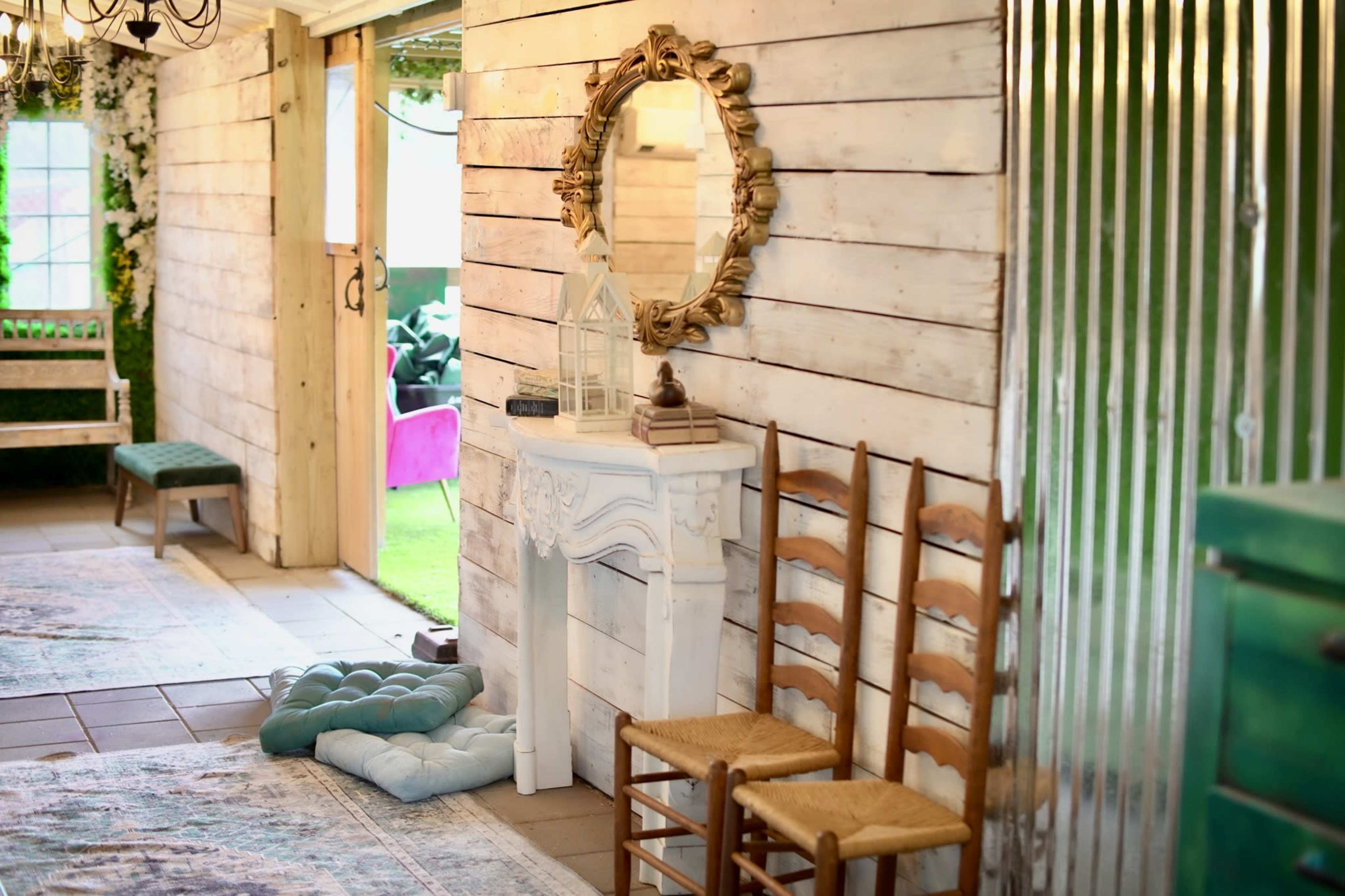 A room with rustic wooden walls, a vintage mirror above a small white console table, and two wooden chairs, alongside a cushioned floor pillow.