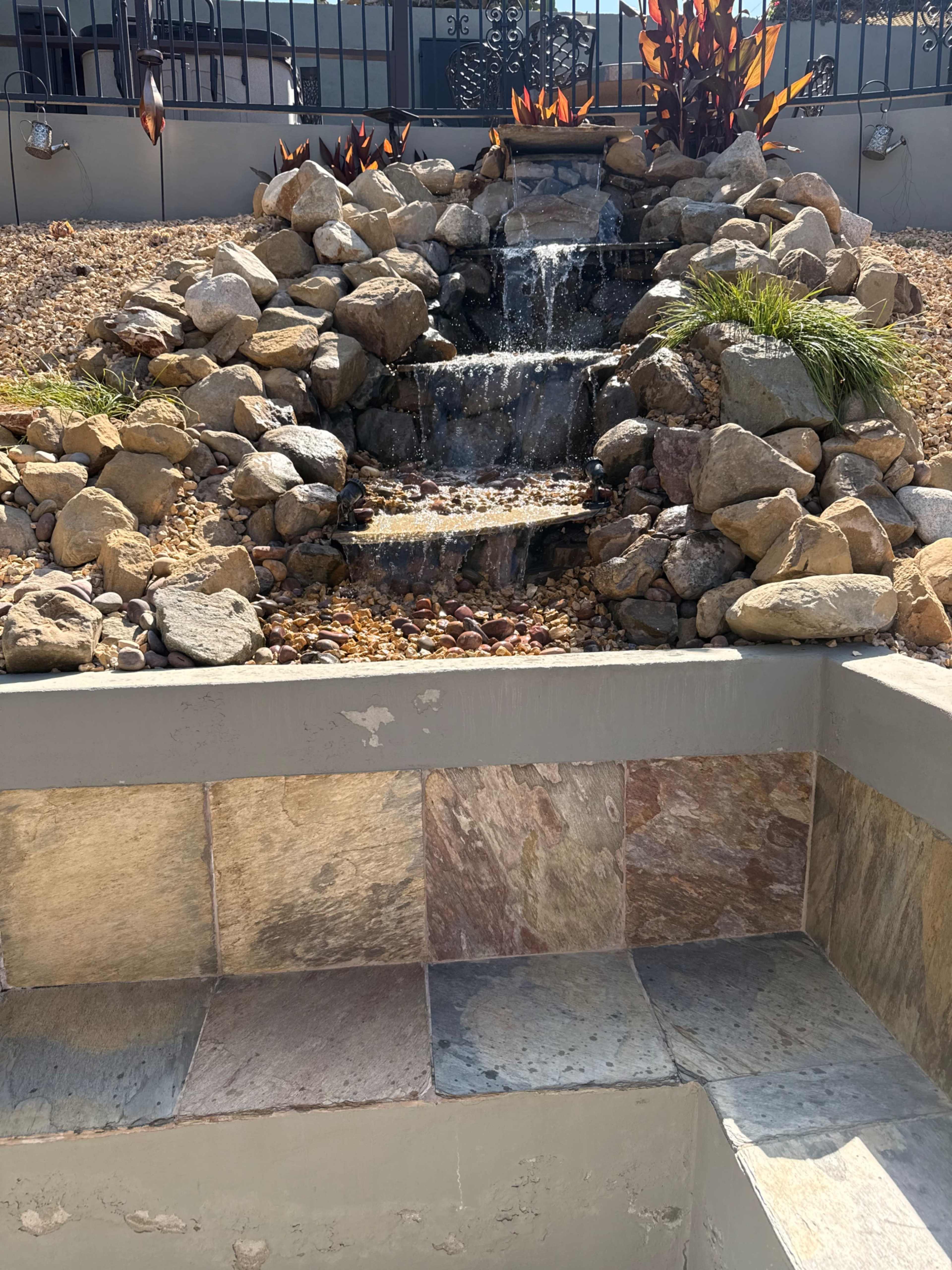 A small waterfall cascades over stones in a landscaped area, with smooth rocks and pebbles arranged around it.