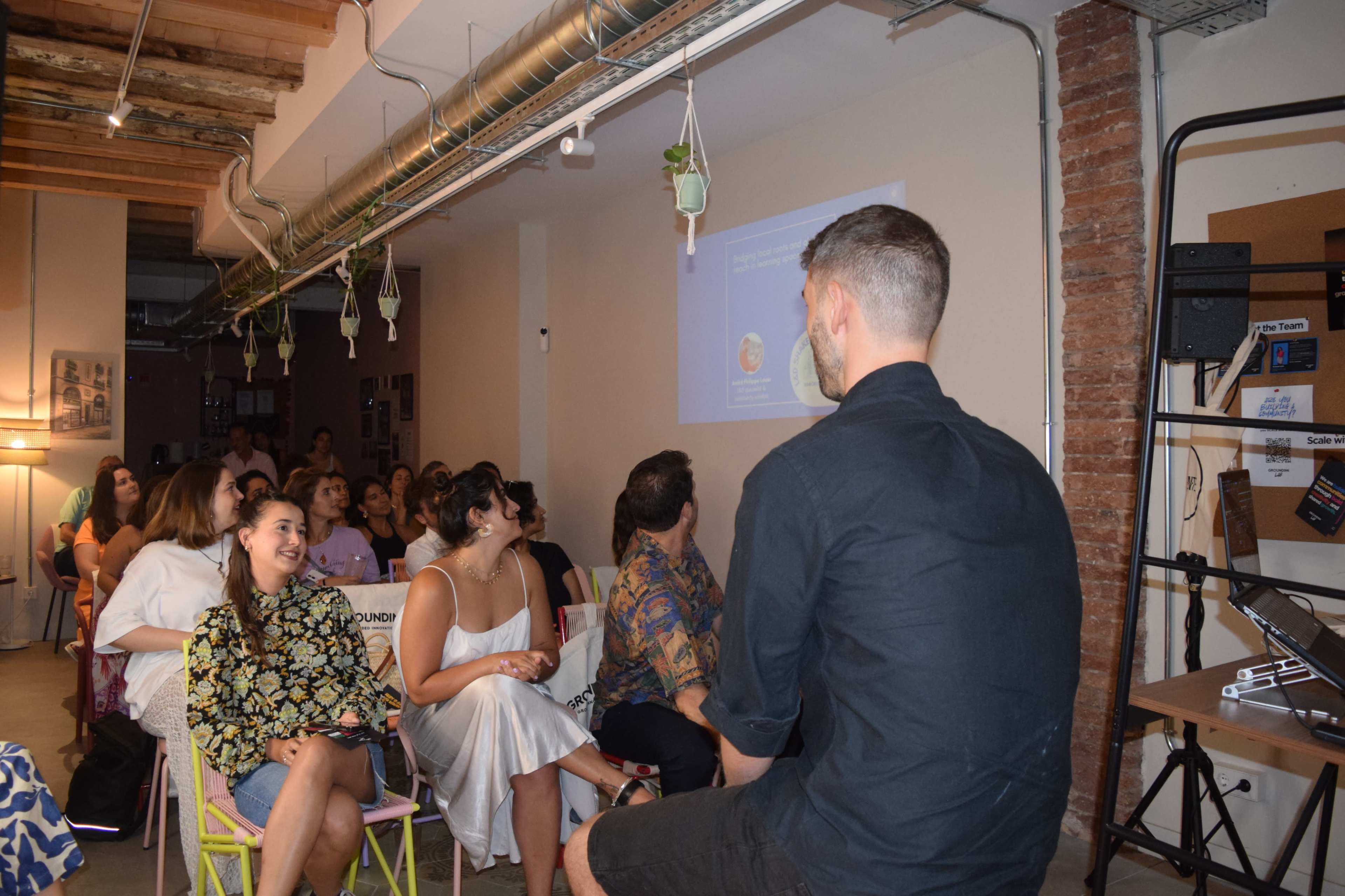 A group of people is seated in a room, watching a presentation displayed on a screen while a speaker is visible in profile at the forefront.