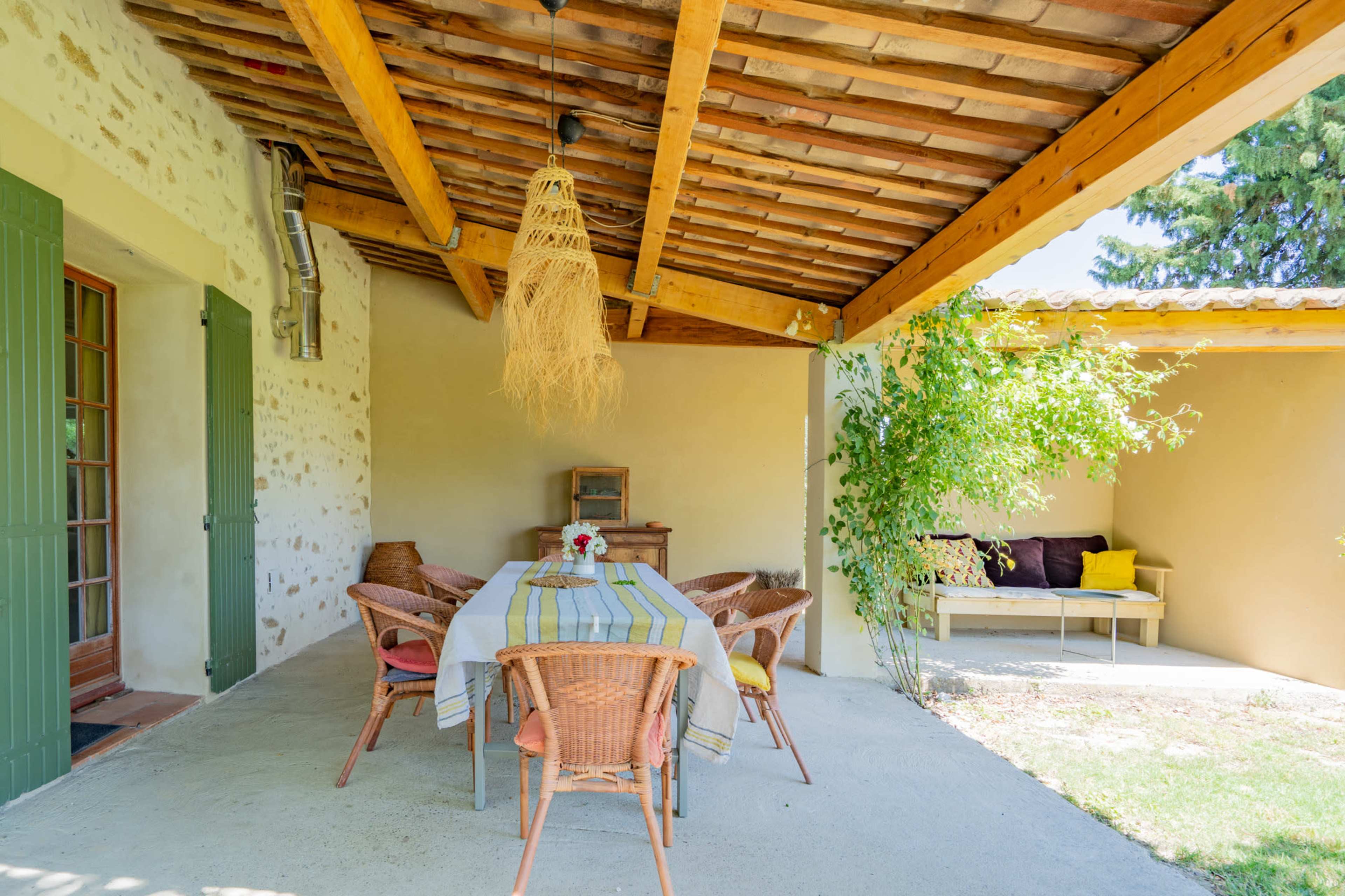 A covered outdoor dining area features a wooden table surrounded by wicker chairs, with a light fixture overhead and greenery along the walls.
