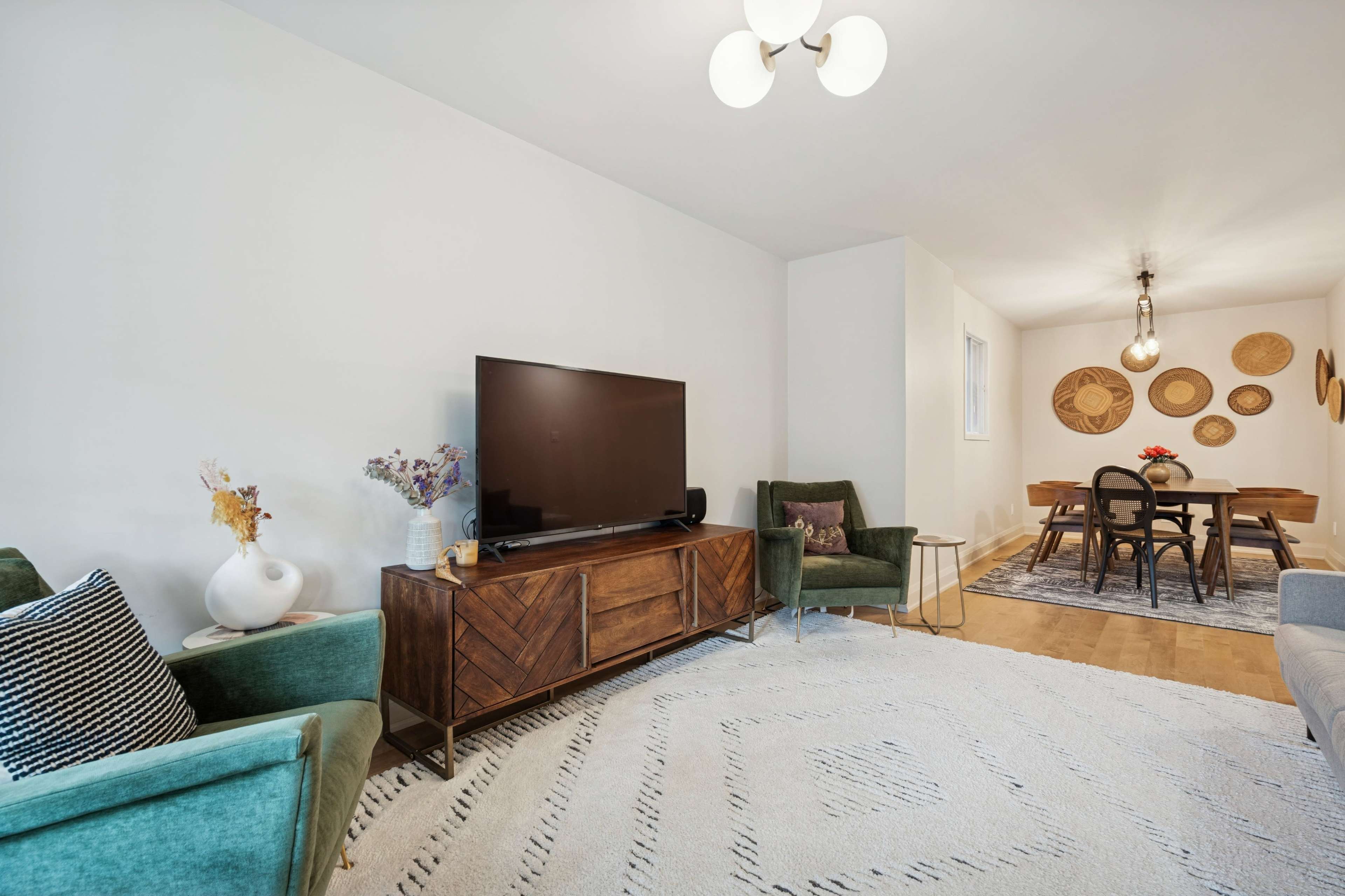 The image features a modern living room with a flat-screen TV on a wooden console, two green armchairs, and a dining area with a table and decorative wall hangings.
