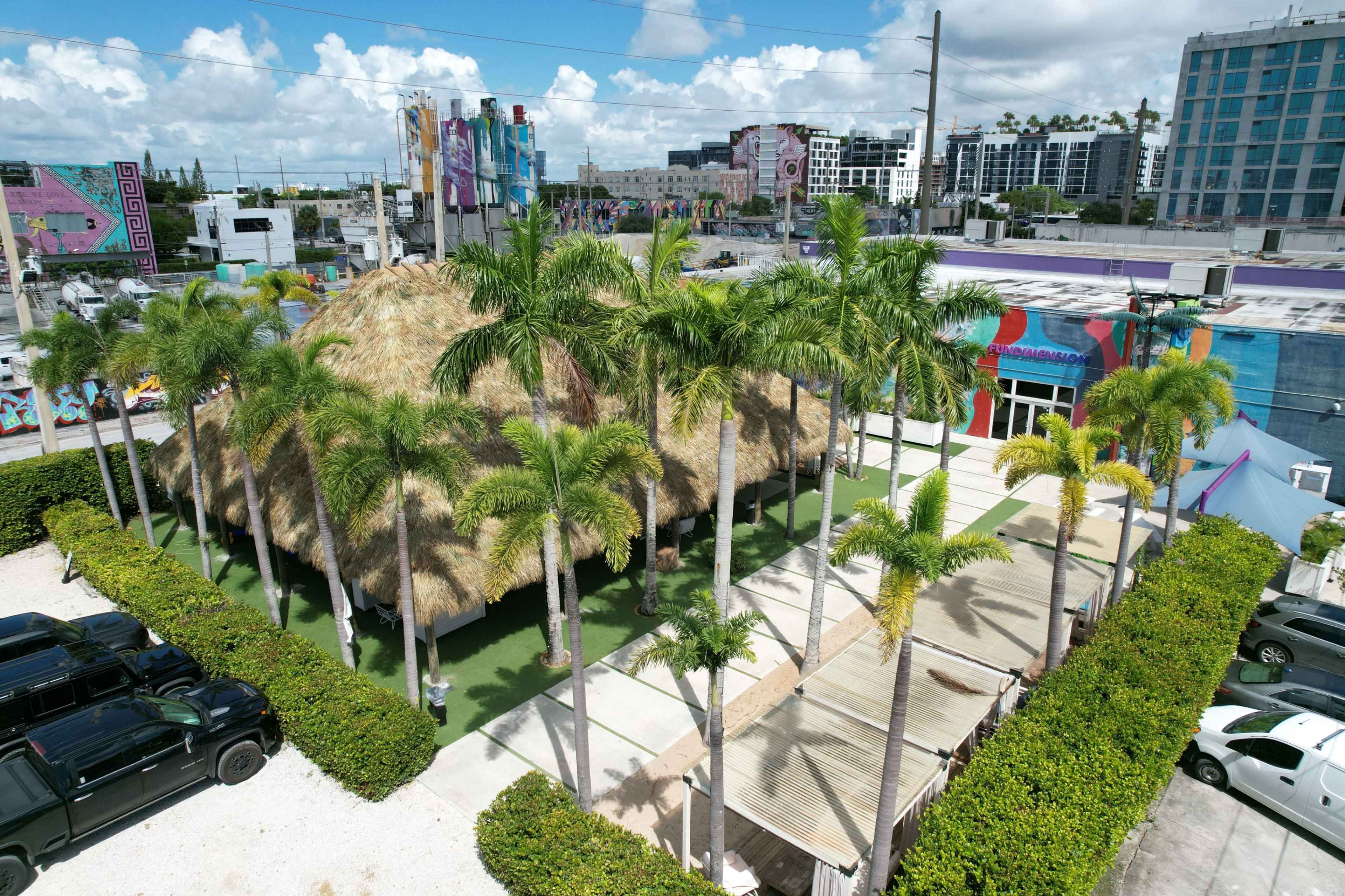 The image shows a large thatched-roof structure surrounded by palm trees, with a paved area and several parked cars nearby.