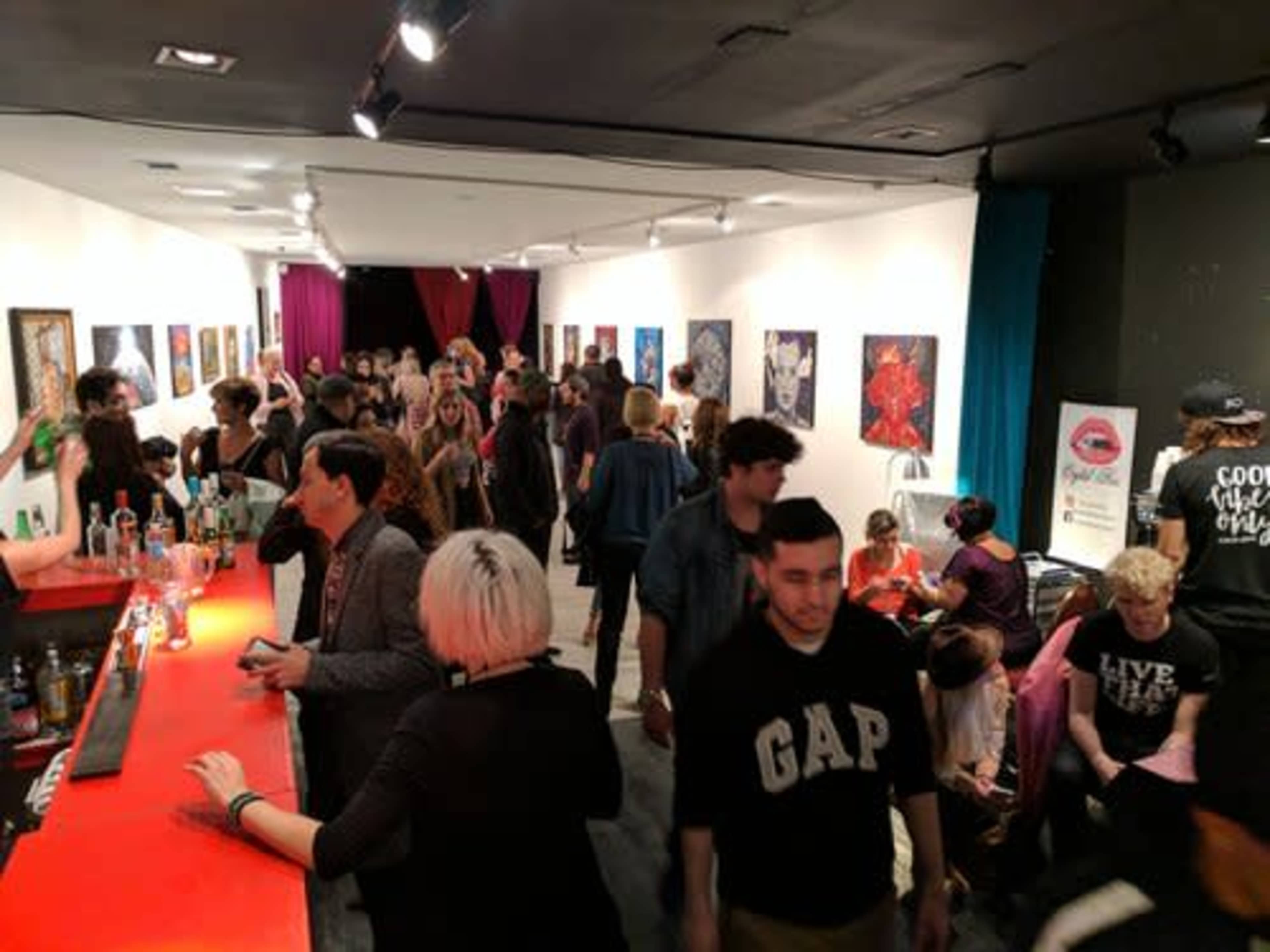 A crowded art gallery features guests mingling and viewing colorful artwork on the walls.
