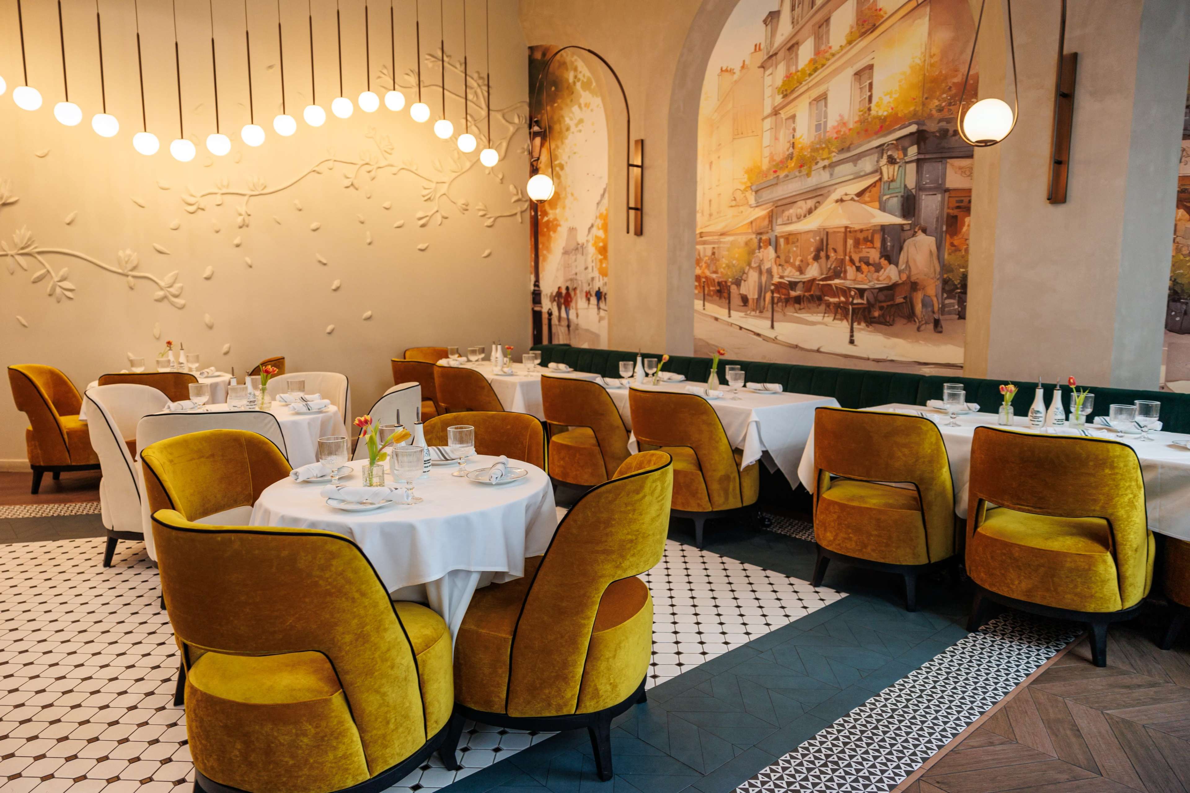 The image shows a modern restaurant interior with round tables covered in white tablecloths and yellow upholstered chairs, illuminated by hanging lights and featuring a mural of a Parisian street scene on one wall.