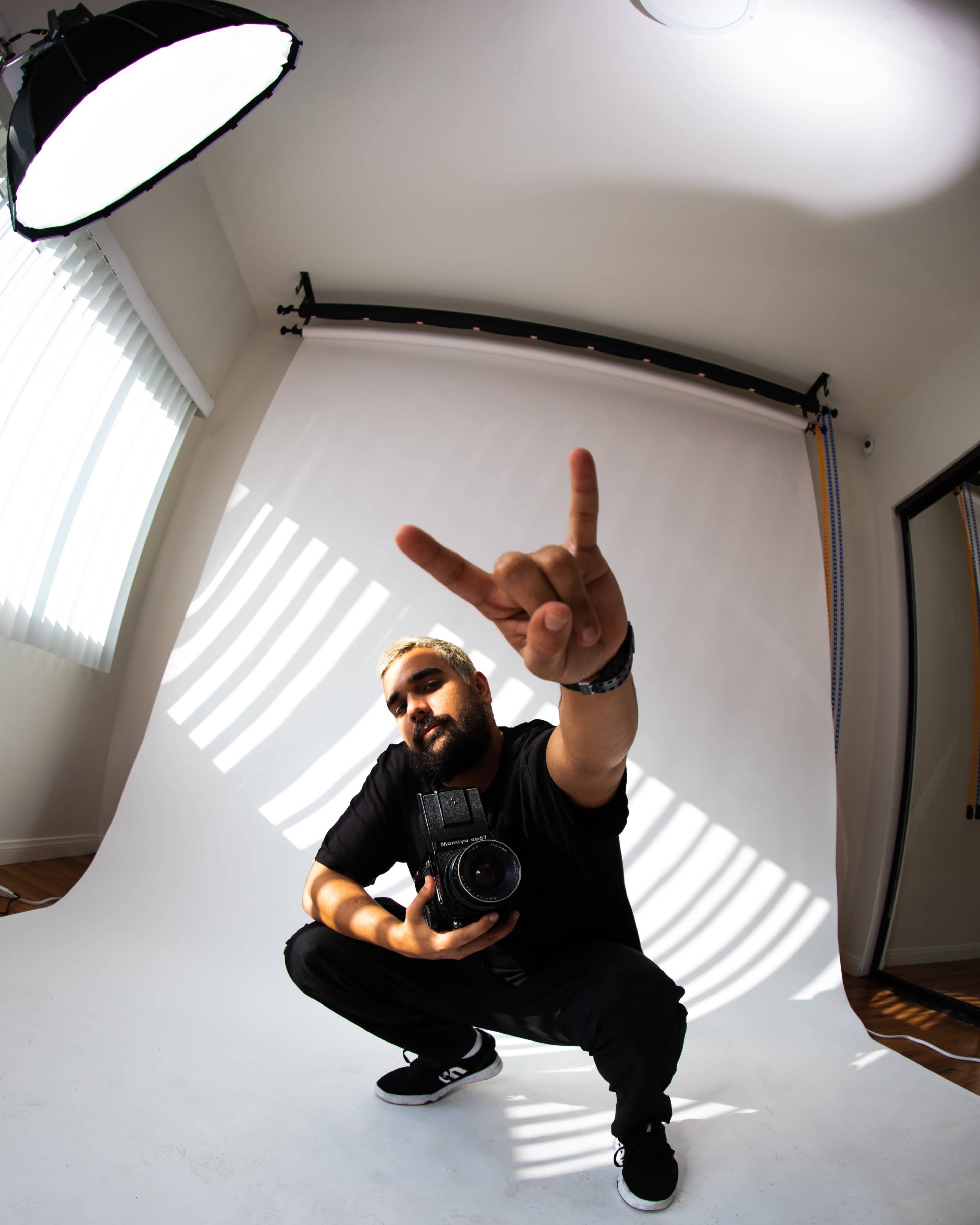A man in a black shirt and cap squats while holding a camera and making a hand gesture against a white backdrop.