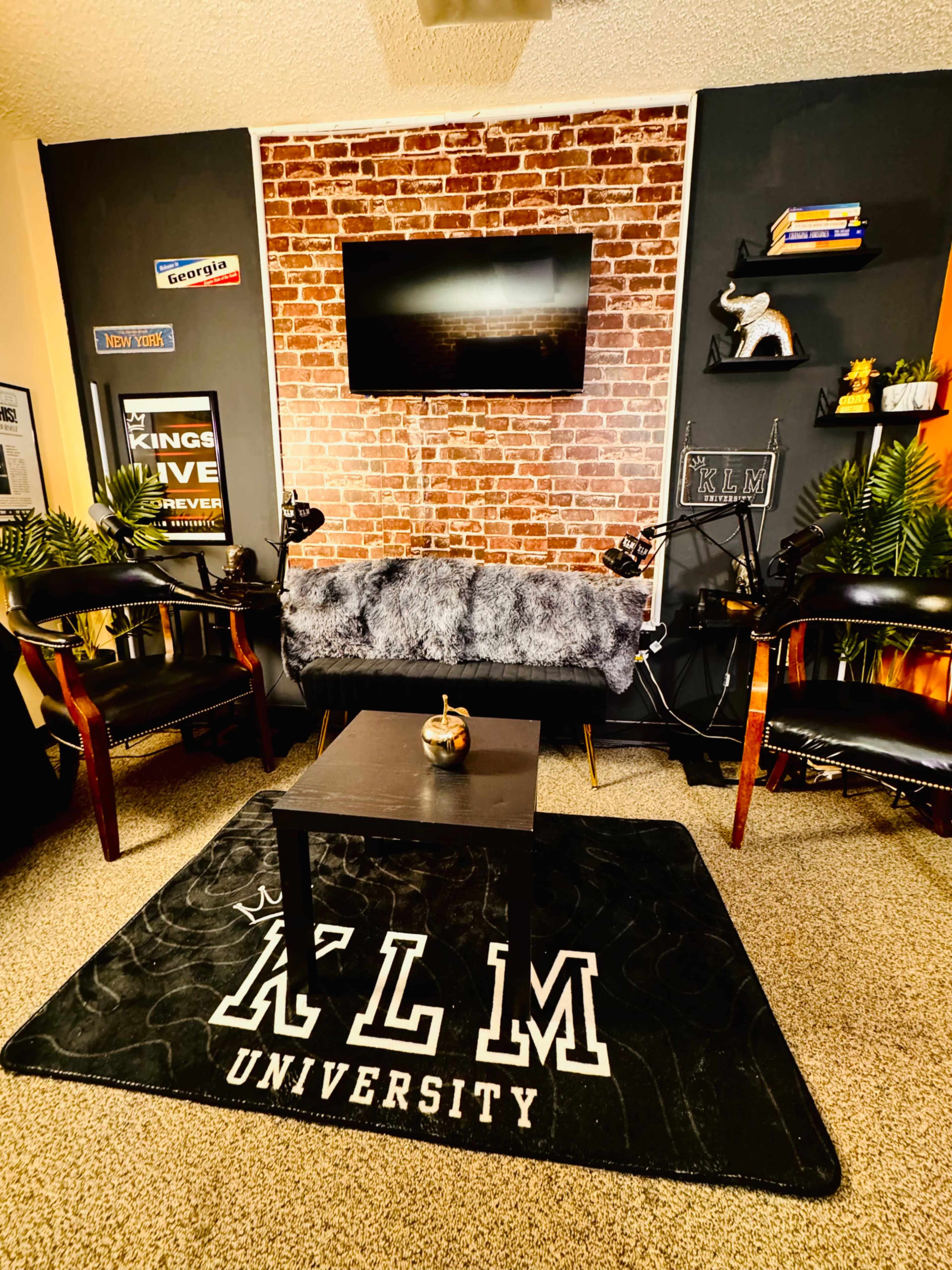 The image shows a cozy living space with a faux brick wall, a television mounted above a gray fur couch, a black coffee table, and two chairs, all arranged on a carpet with "KLM UNIVERSITY" printed on it.