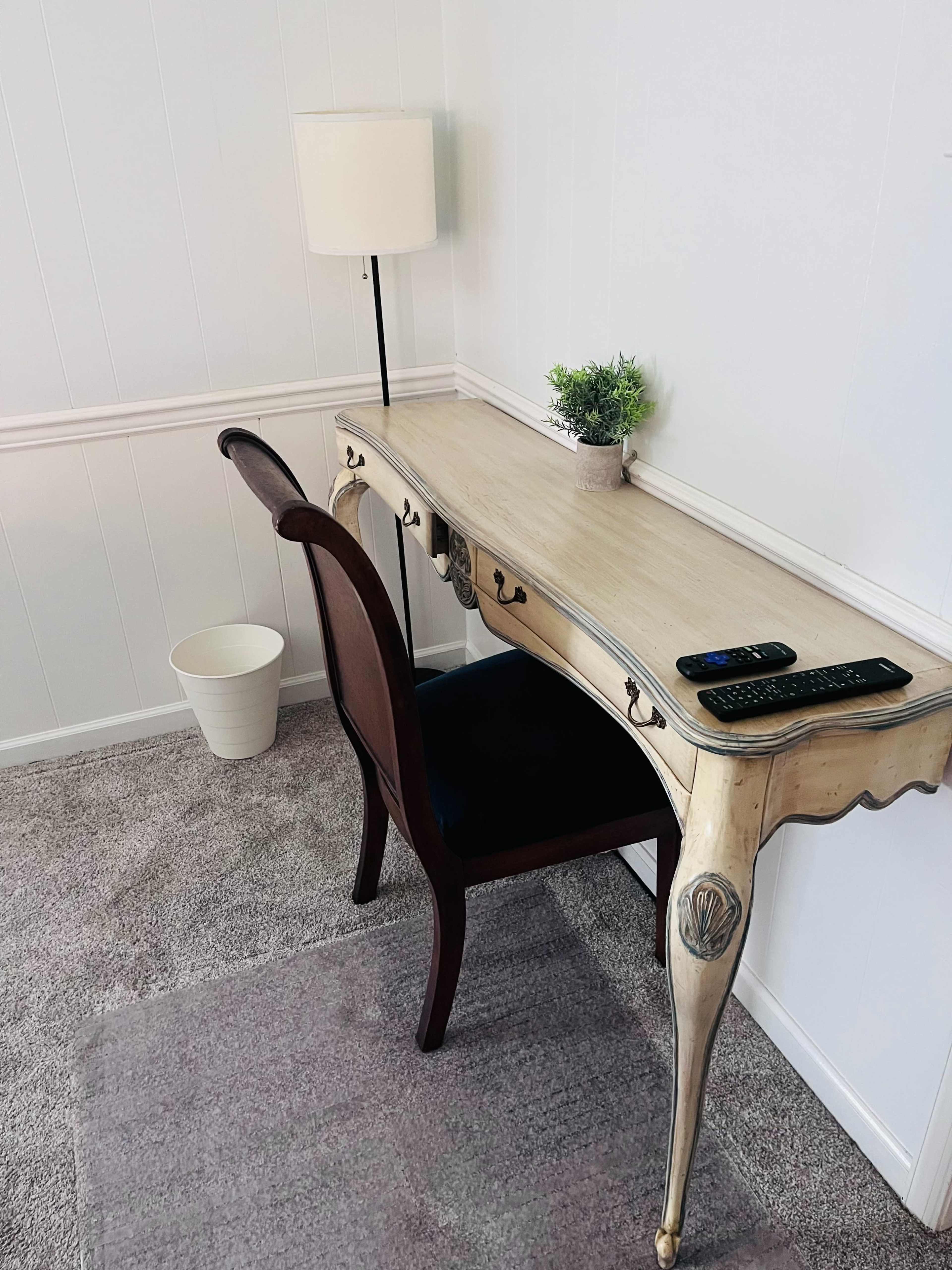 A wooden desk with a chair is positioned against a white wall, accompanied by a lamp and a small potted plant.