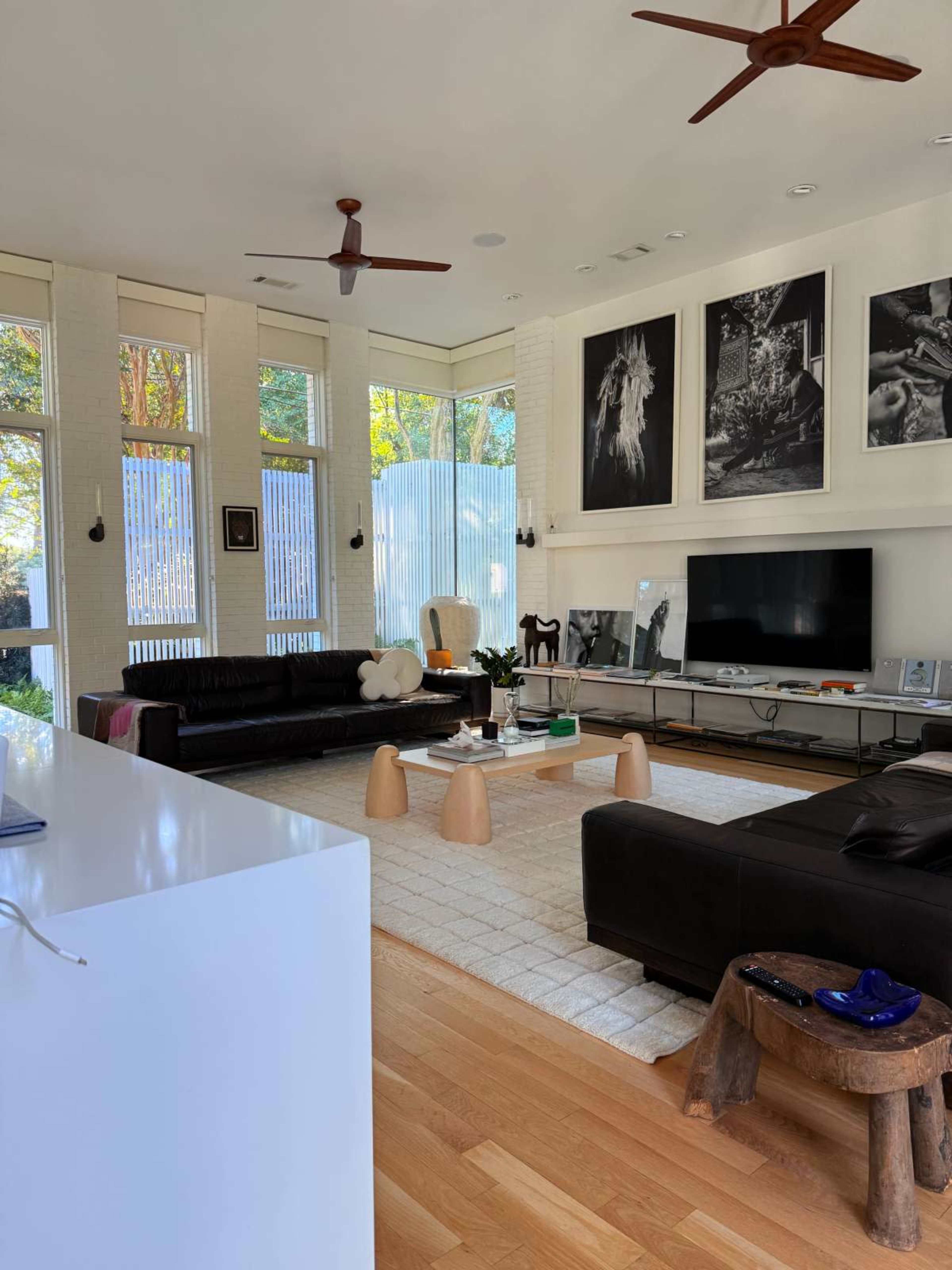 The image shows a modern living room with large windows, two black sofas, a wooden coffee table, and several pieces of wall art.
