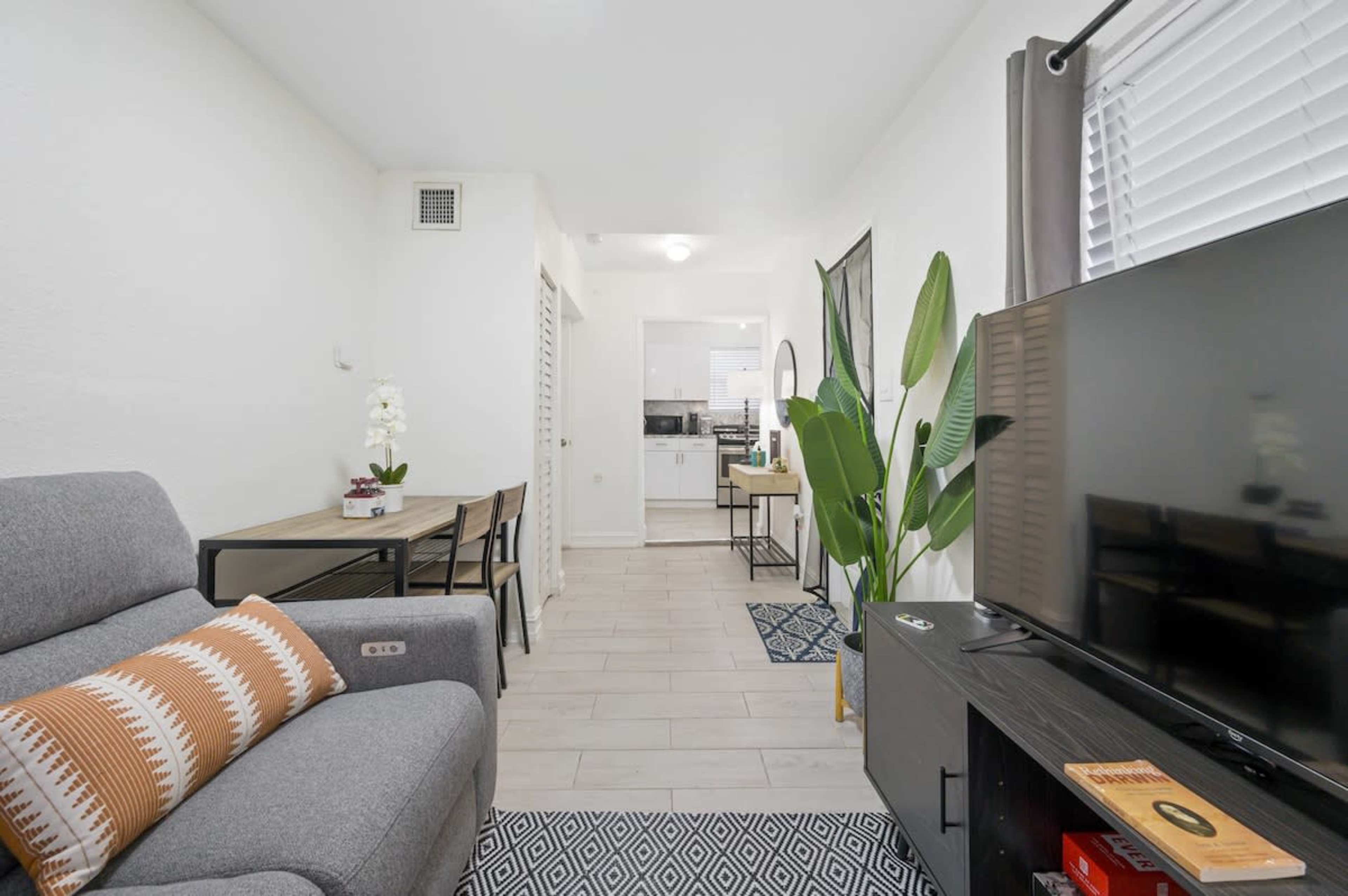 A modern living space with a gray sofa, a dining area, and a television set, featuring large windows and indoor plants.