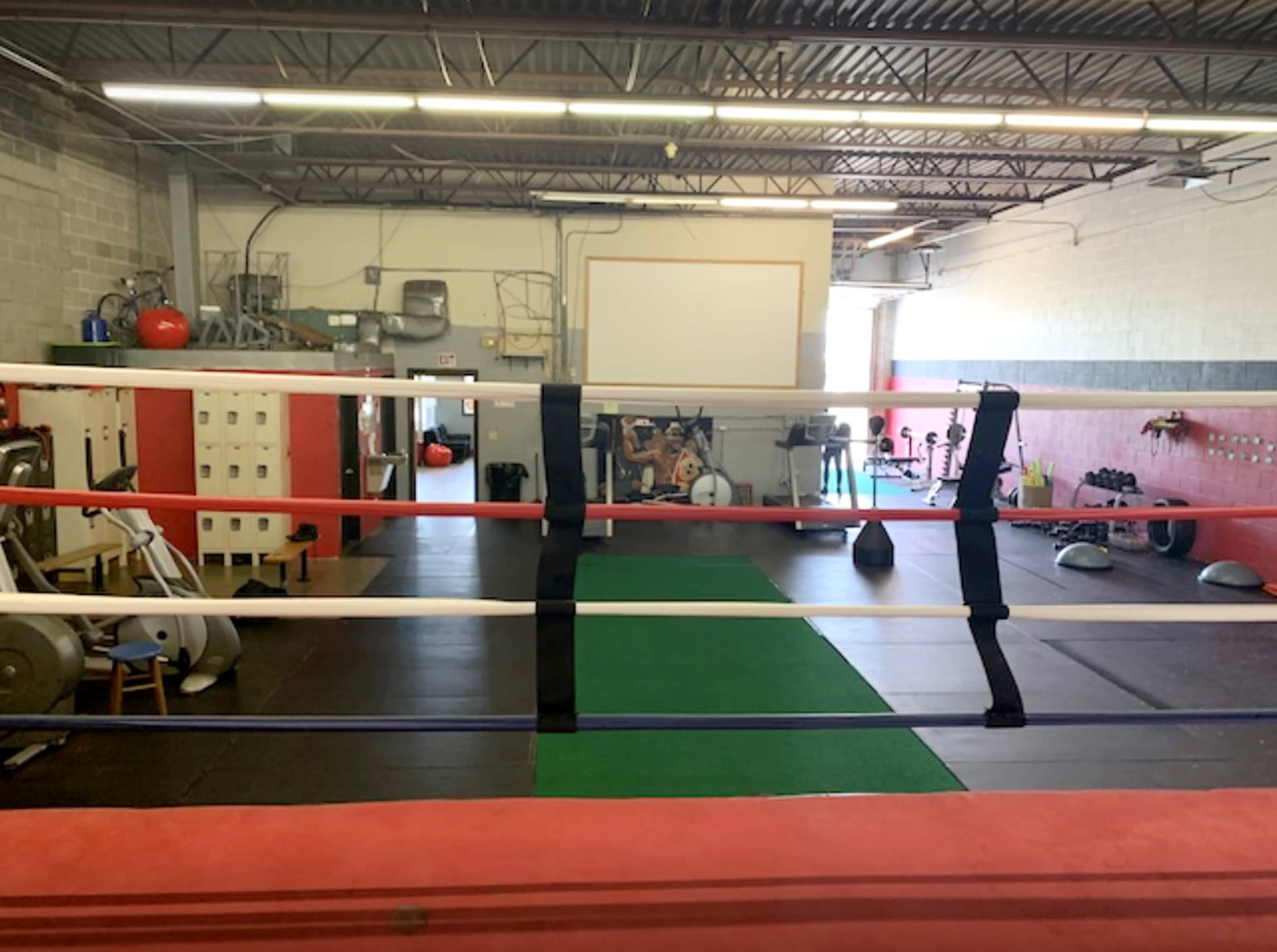 A boxing gym with a red and white boxing ring, equipment stored nearby, and a workout area featuring mats and weights.