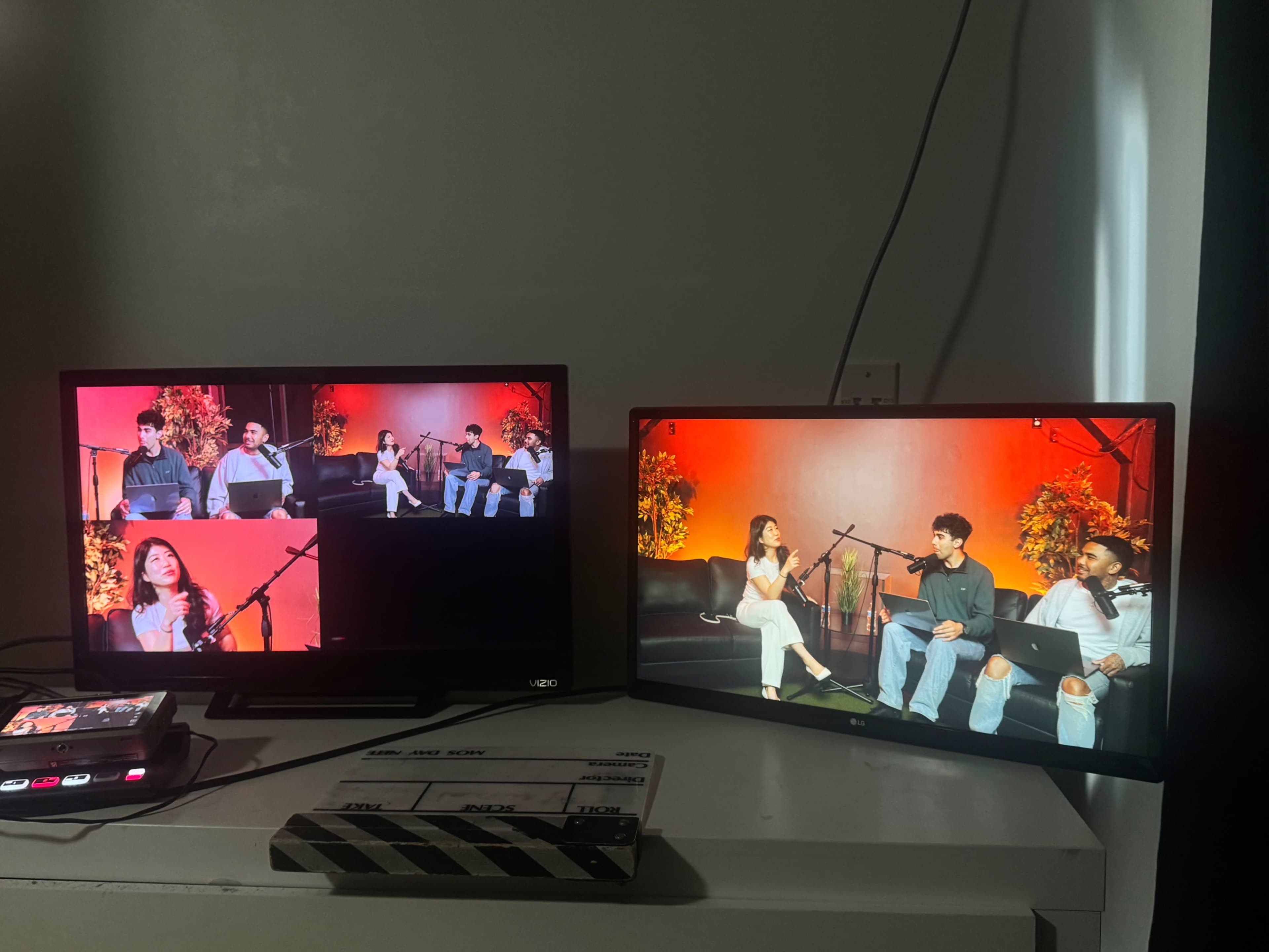 Two televisions display a recording of a panel discussion featuring three individuals, with one woman and two men, set against a backdrop of orange lighting and plants.