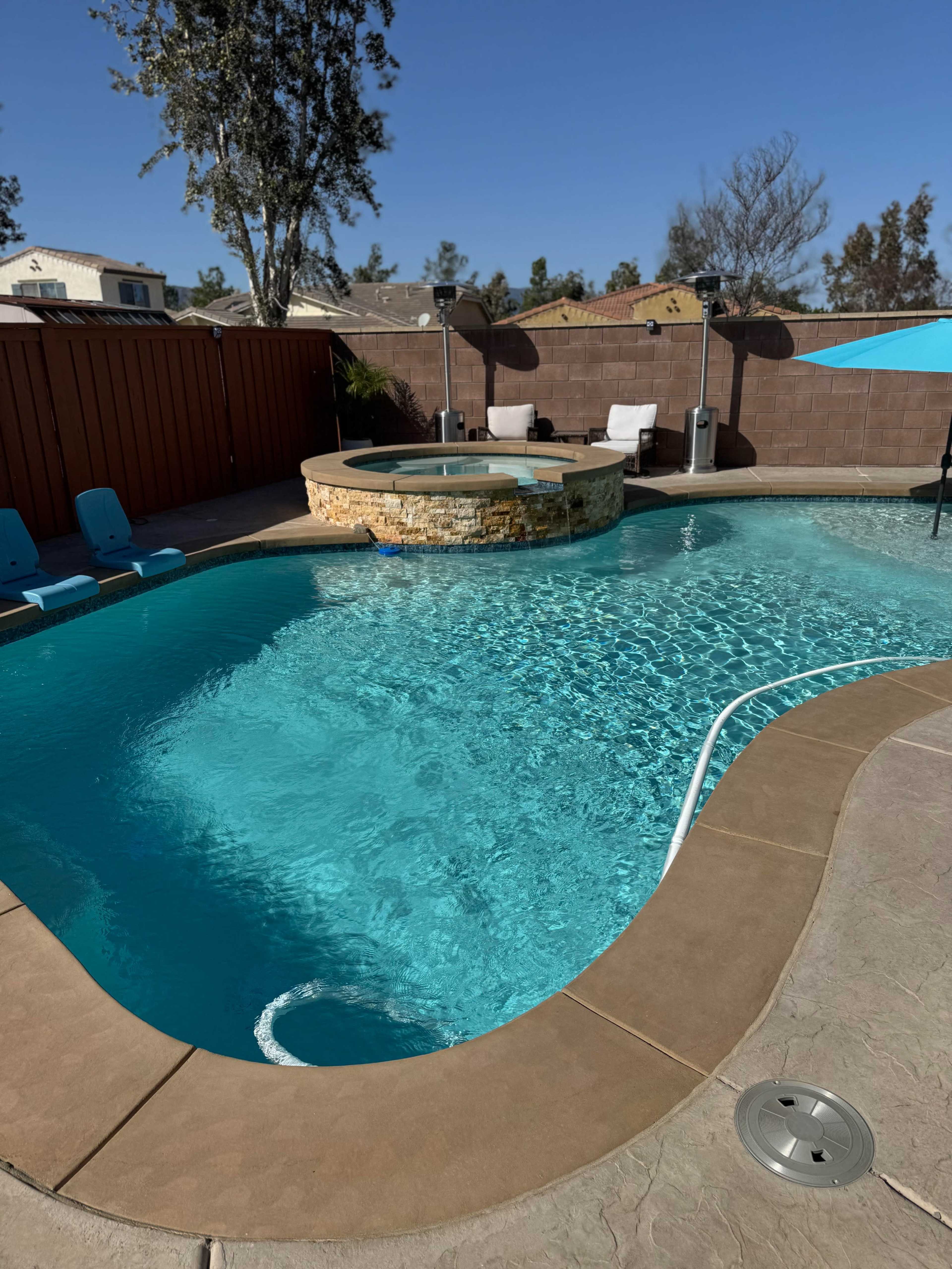 The image shows a backyard swimming pool with a spa area, surrounded by a wooden fence and patio furniture under a clear blue sky.