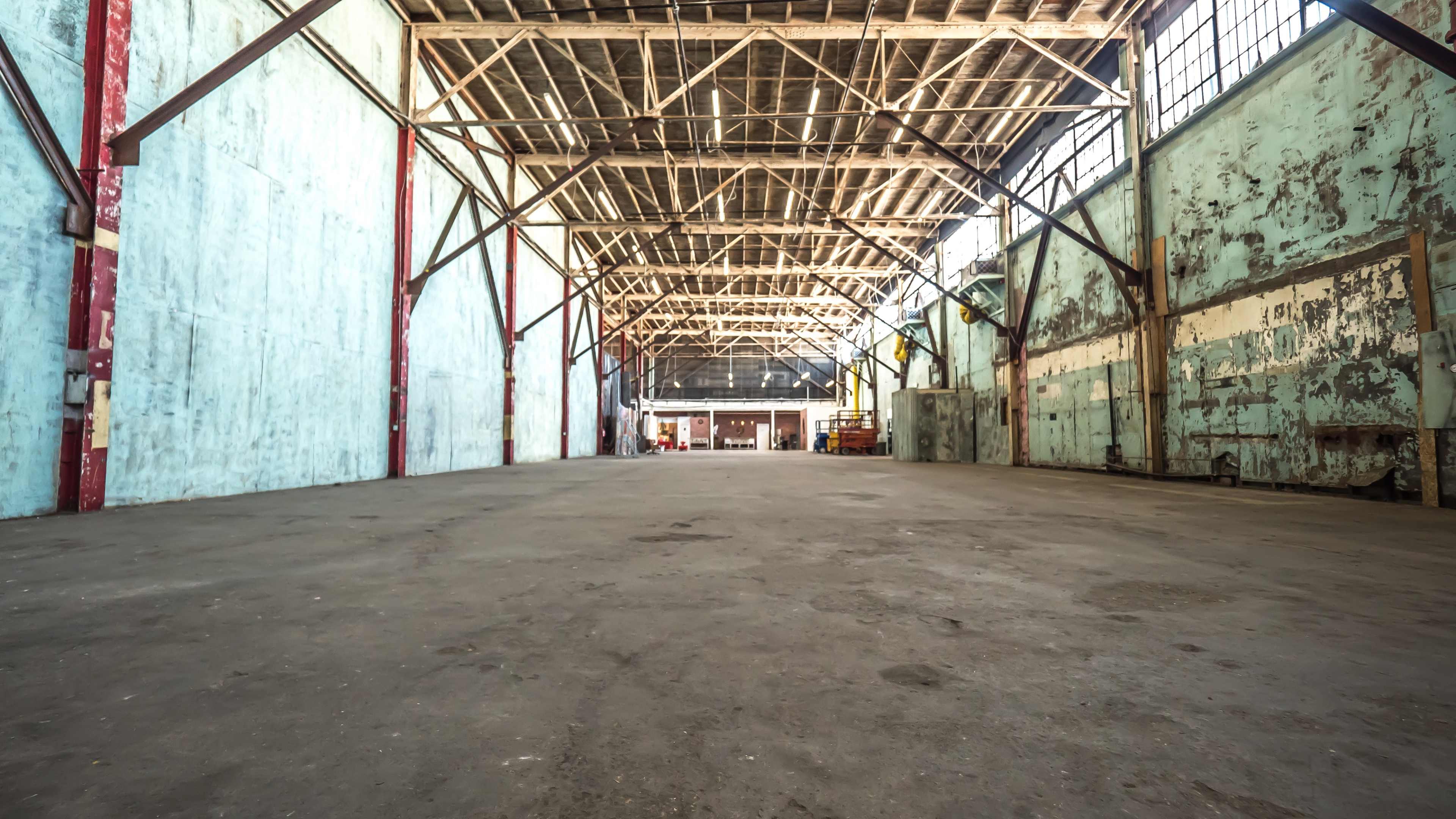 The image shows an empty warehouse with high ceilings, exposed wooden beams, and worn concrete flooring.