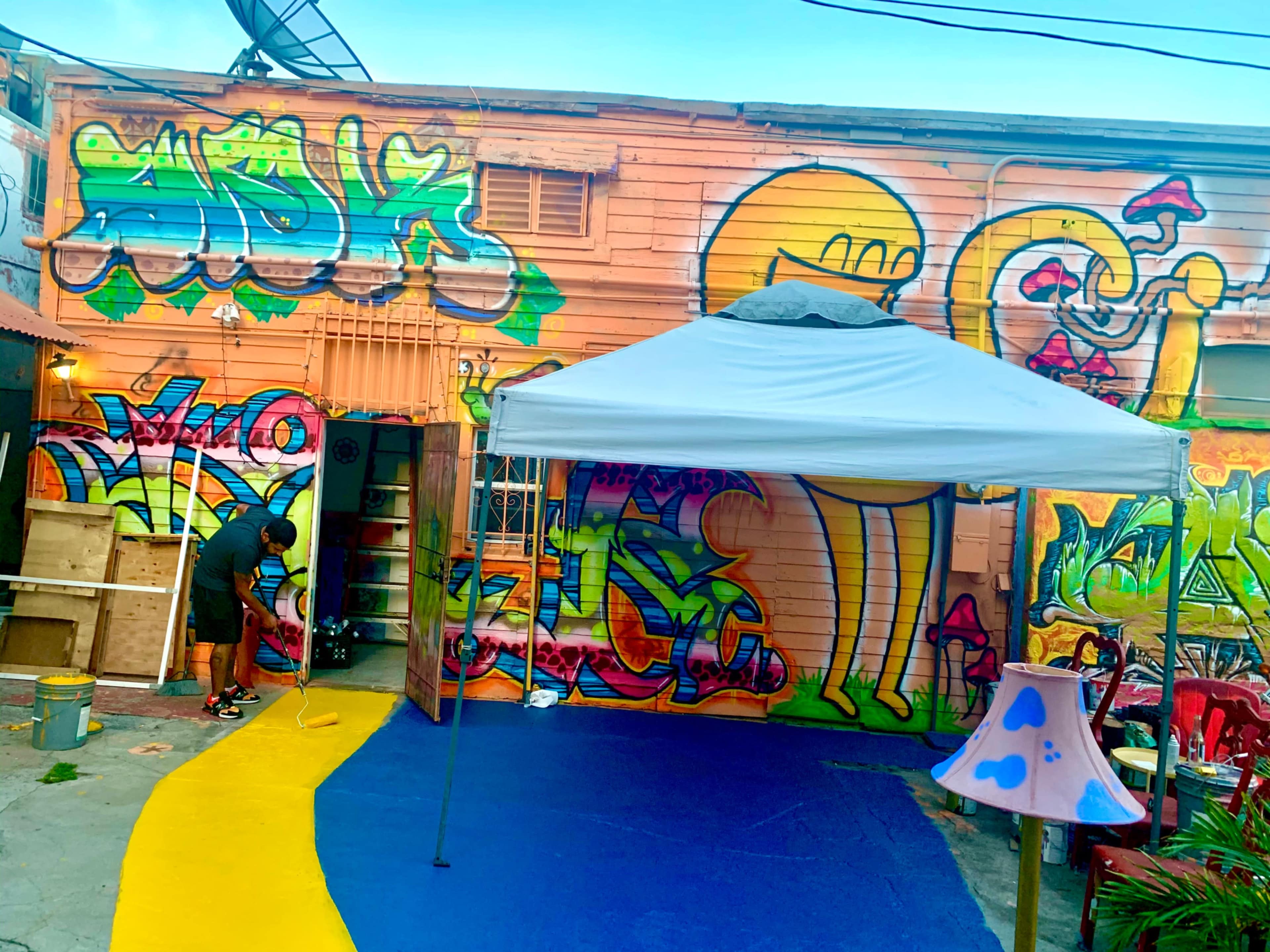 A person is painting a colorful mural on a wall of a building adorned with graffiti, while a tent covers a blue area with yellow accents in the foreground.