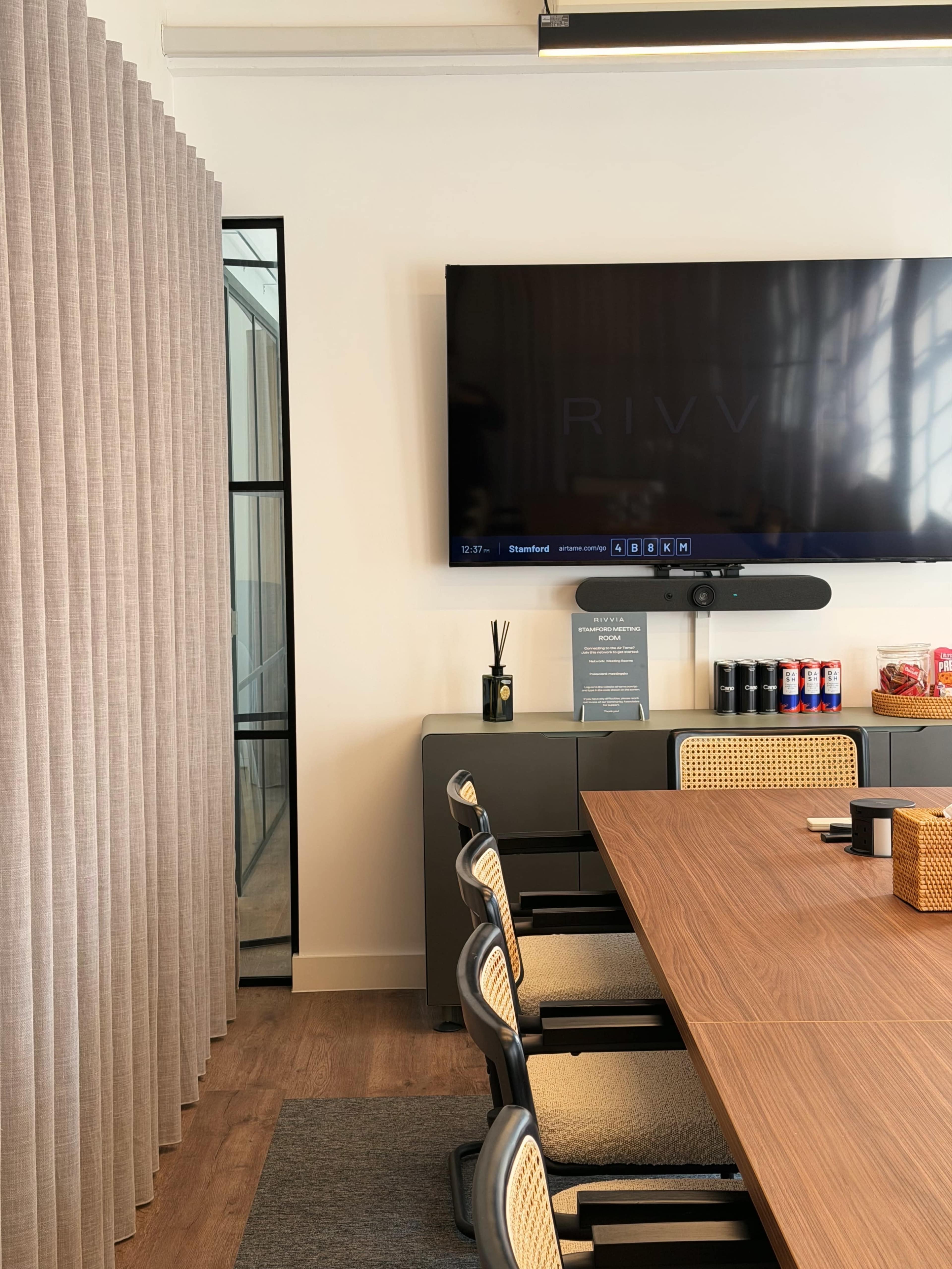 The image shows a modern conference room with a large table, a wall-mounted screen, and a curtain separating the space from an adjacent area.