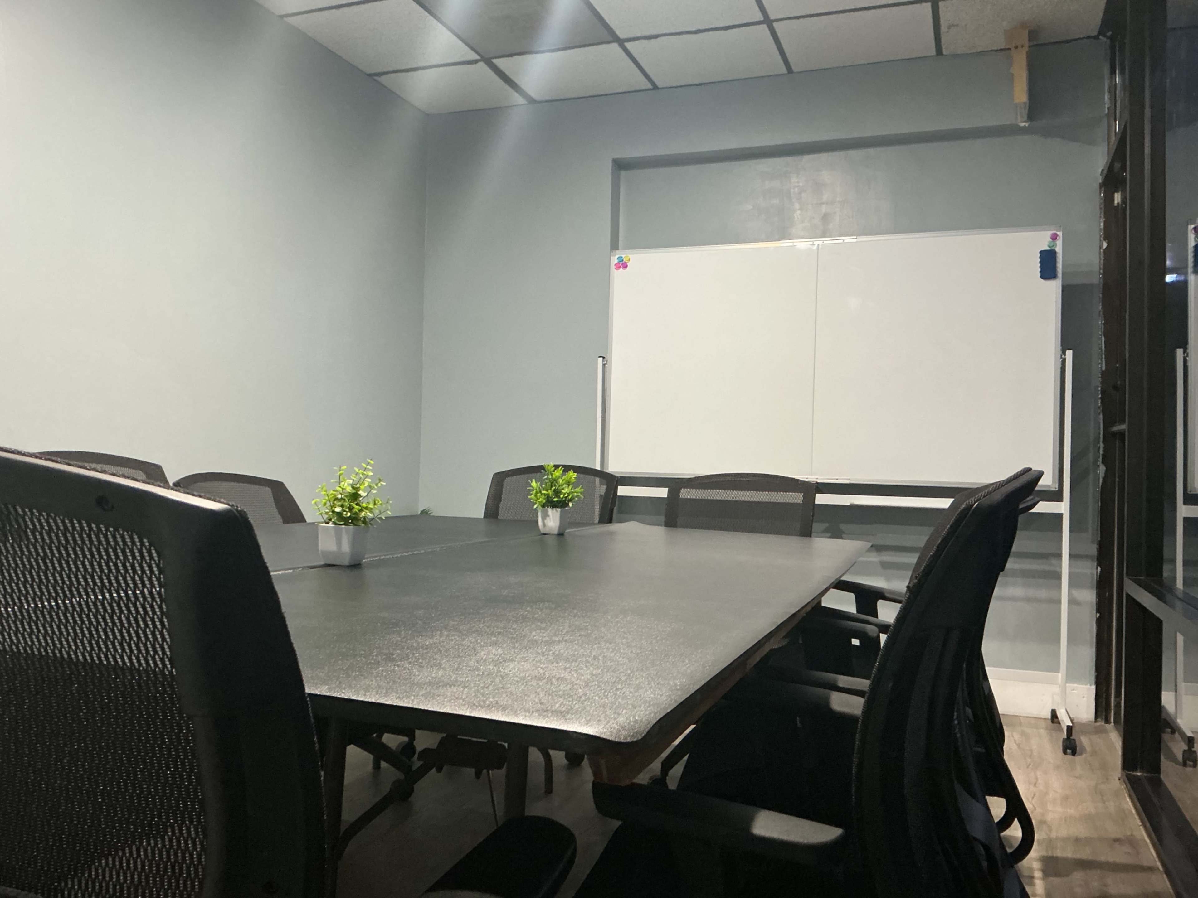 The image shows a conference room with a large dark table, several black chairs, and a whiteboard mounted on a gray wall.