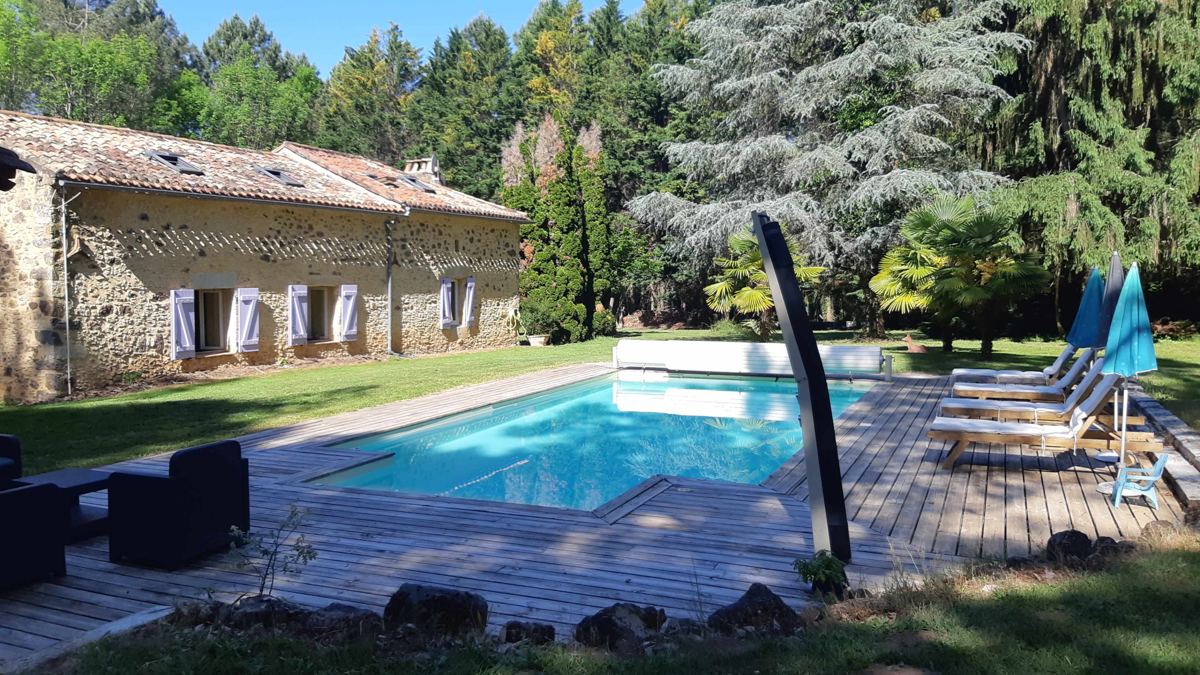 The image shows a stone house next to a swimming pool surrounded by a wooden deck and green trees.