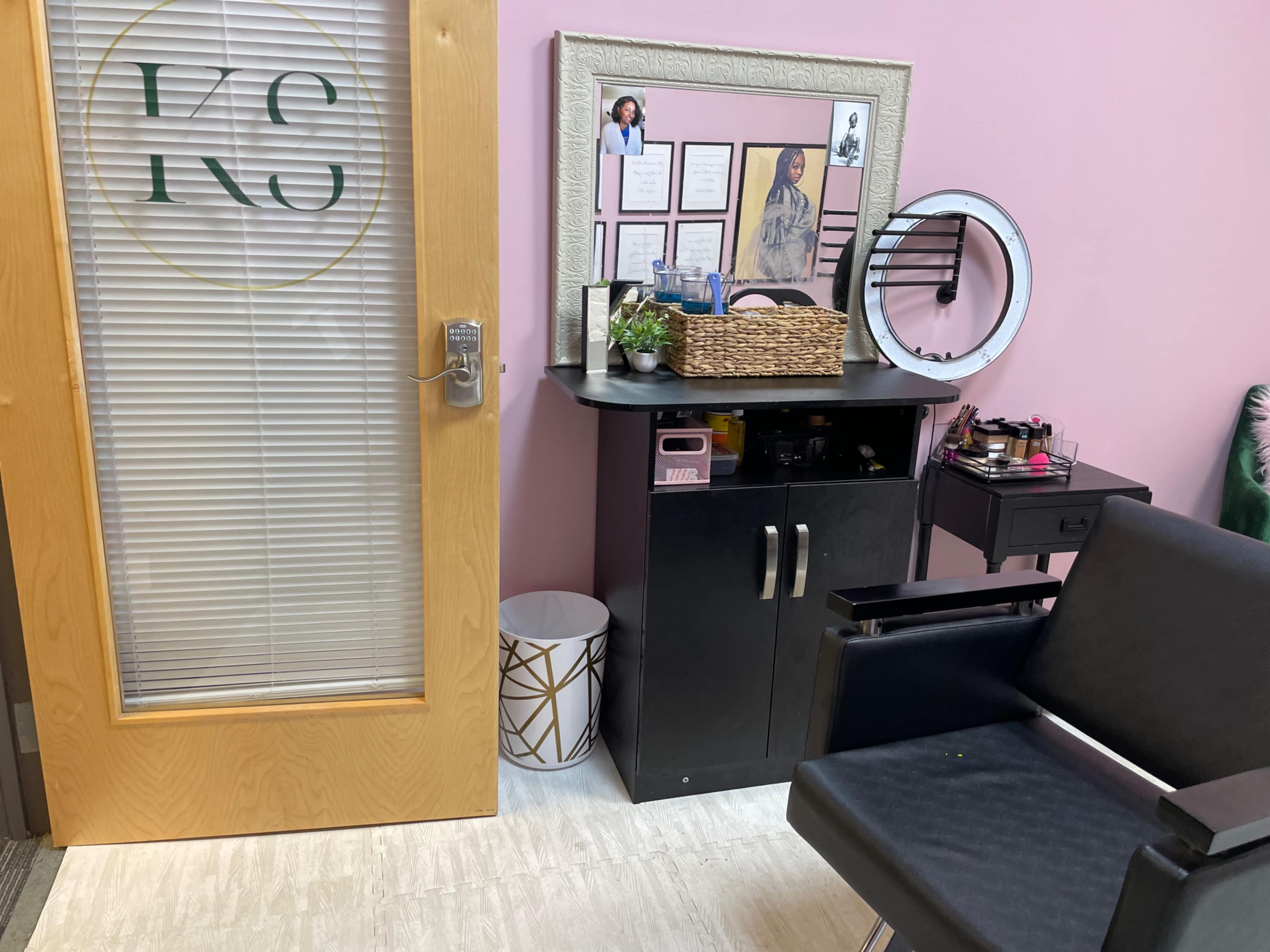 A beauty treatment room featuring a wooden door with the initials "KS," a black vanity with various beauty products, and a comfortable chair next to a patterned wastebasket.