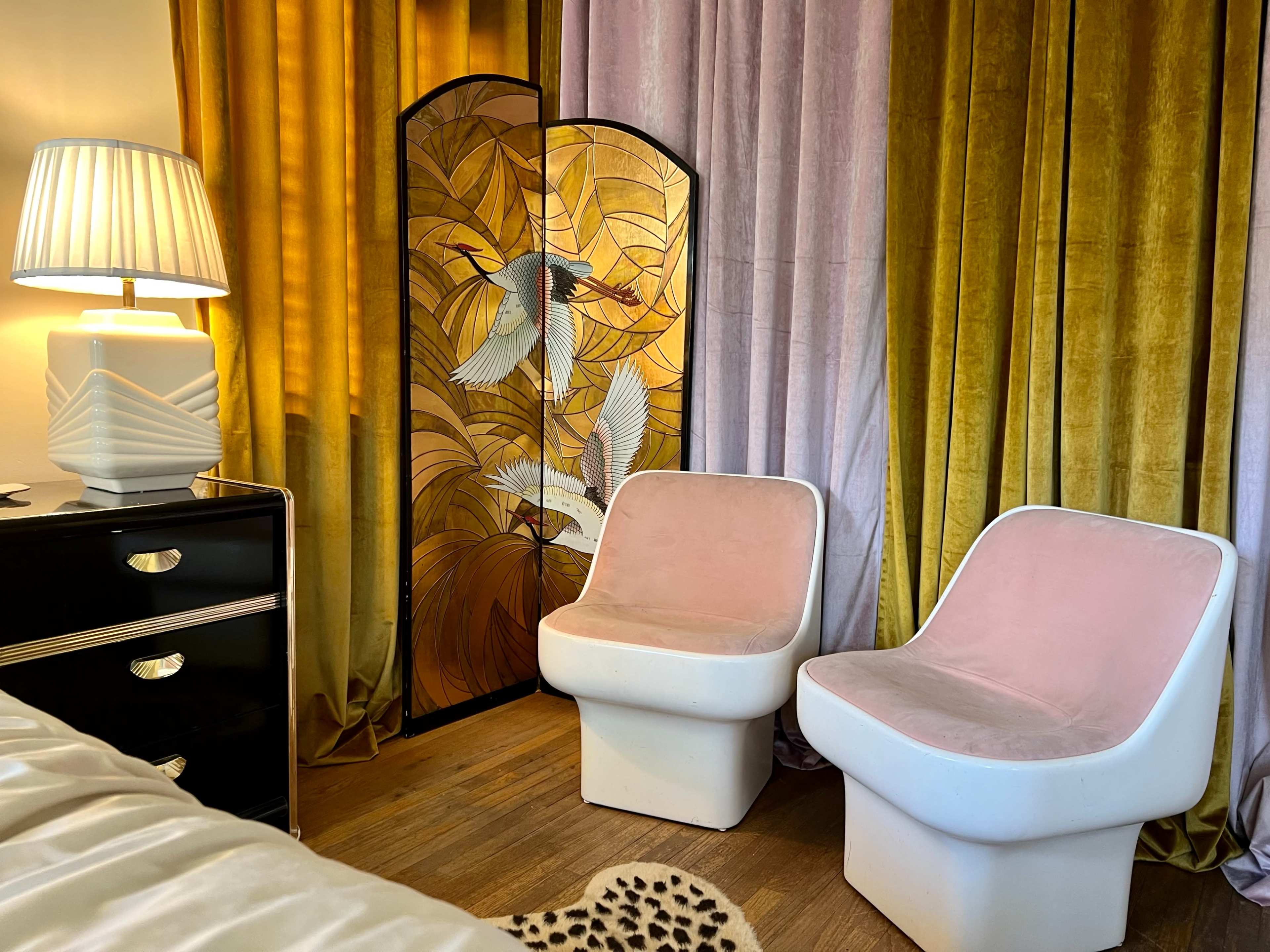 A room with two pink chairs, a lamp, a black nightstand, and a decorative folding screen featuring bird designs, all set against curtains of gold and purple hues.