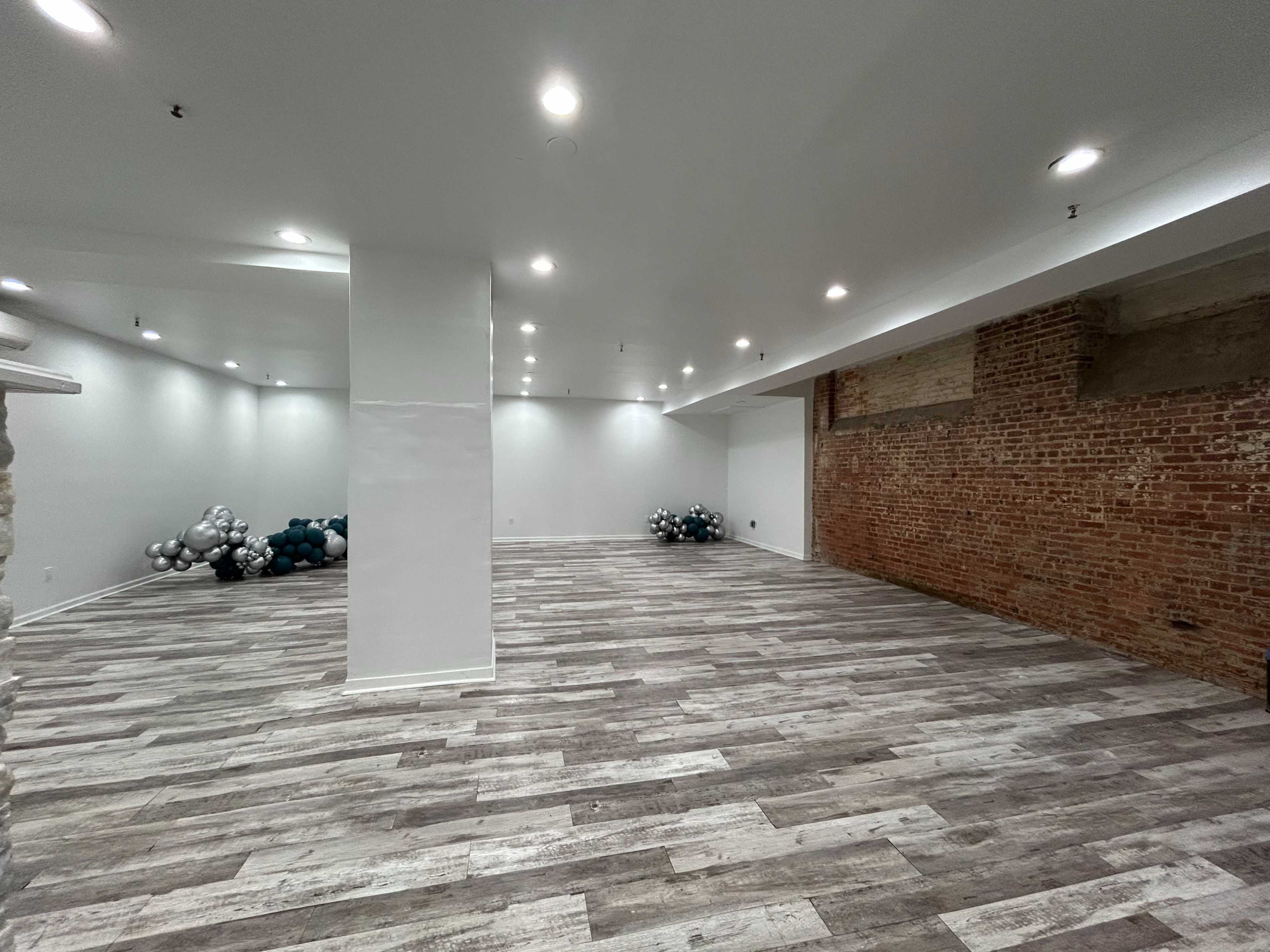 The image shows a spacious, empty room with exposed brick walls, wooden flooring, and clusters of balloons in the corners.