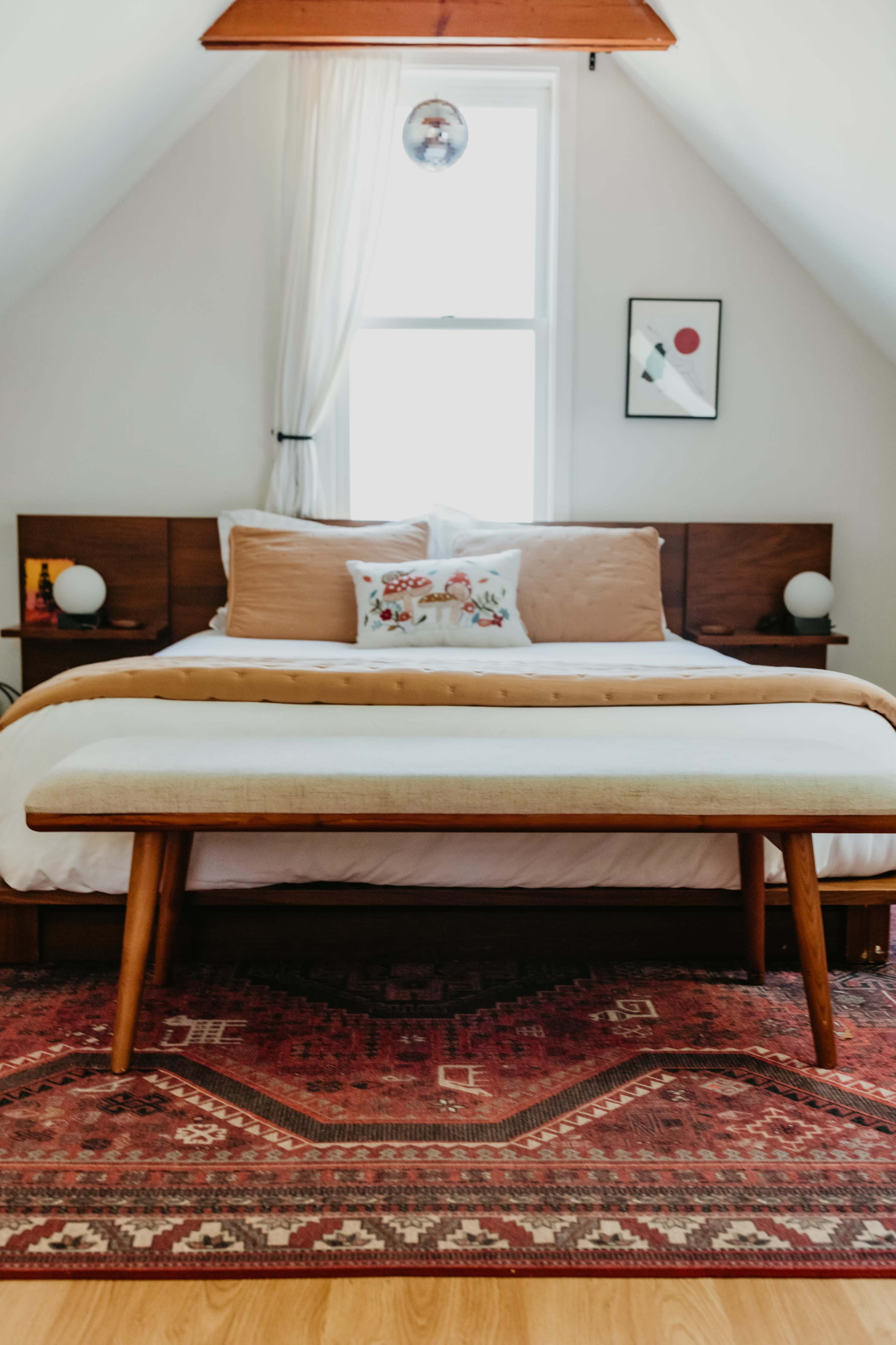 A neatly arranged bedroom features a bed with a decorative throw and a bench at the foot, complemented by a colorful rug and framed artwork on the walls.