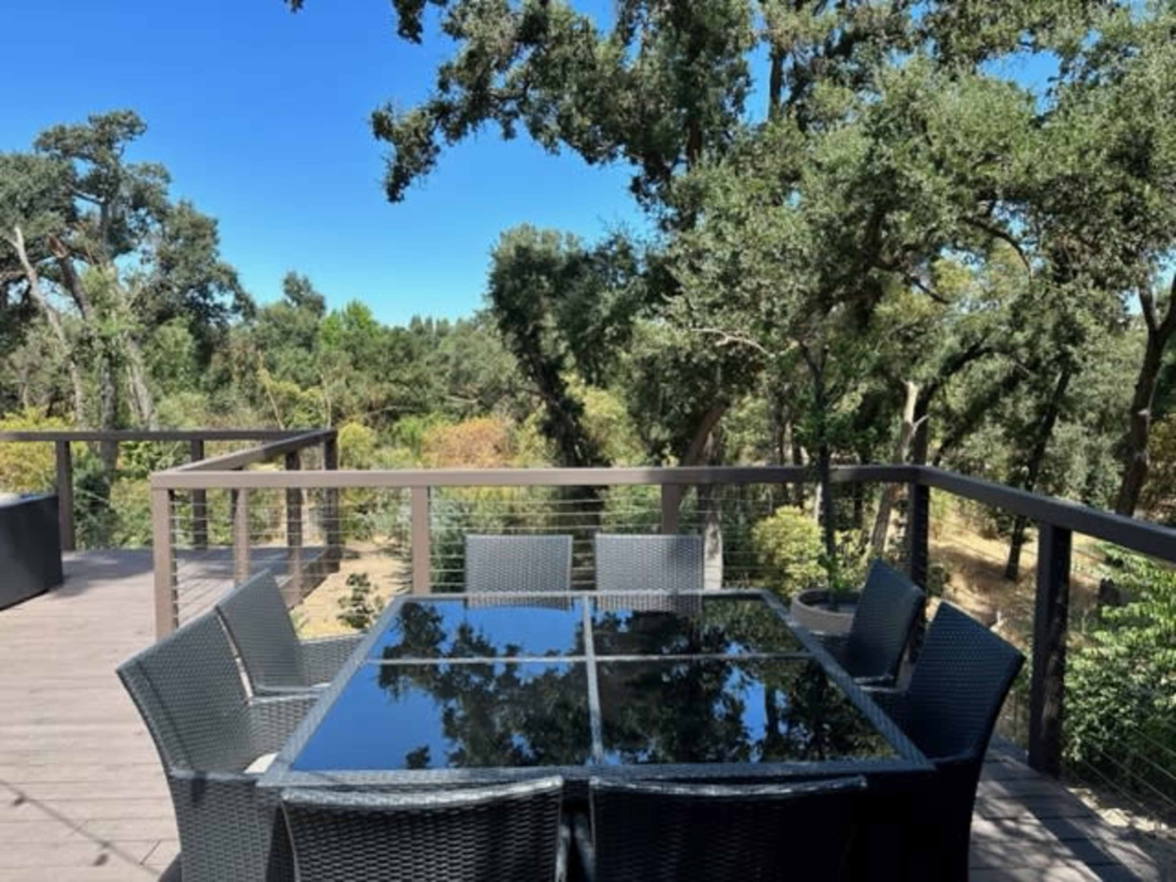 A rectangular glass-top table with six chairs is set on a wooden deck overlooking a lush landscape of trees.