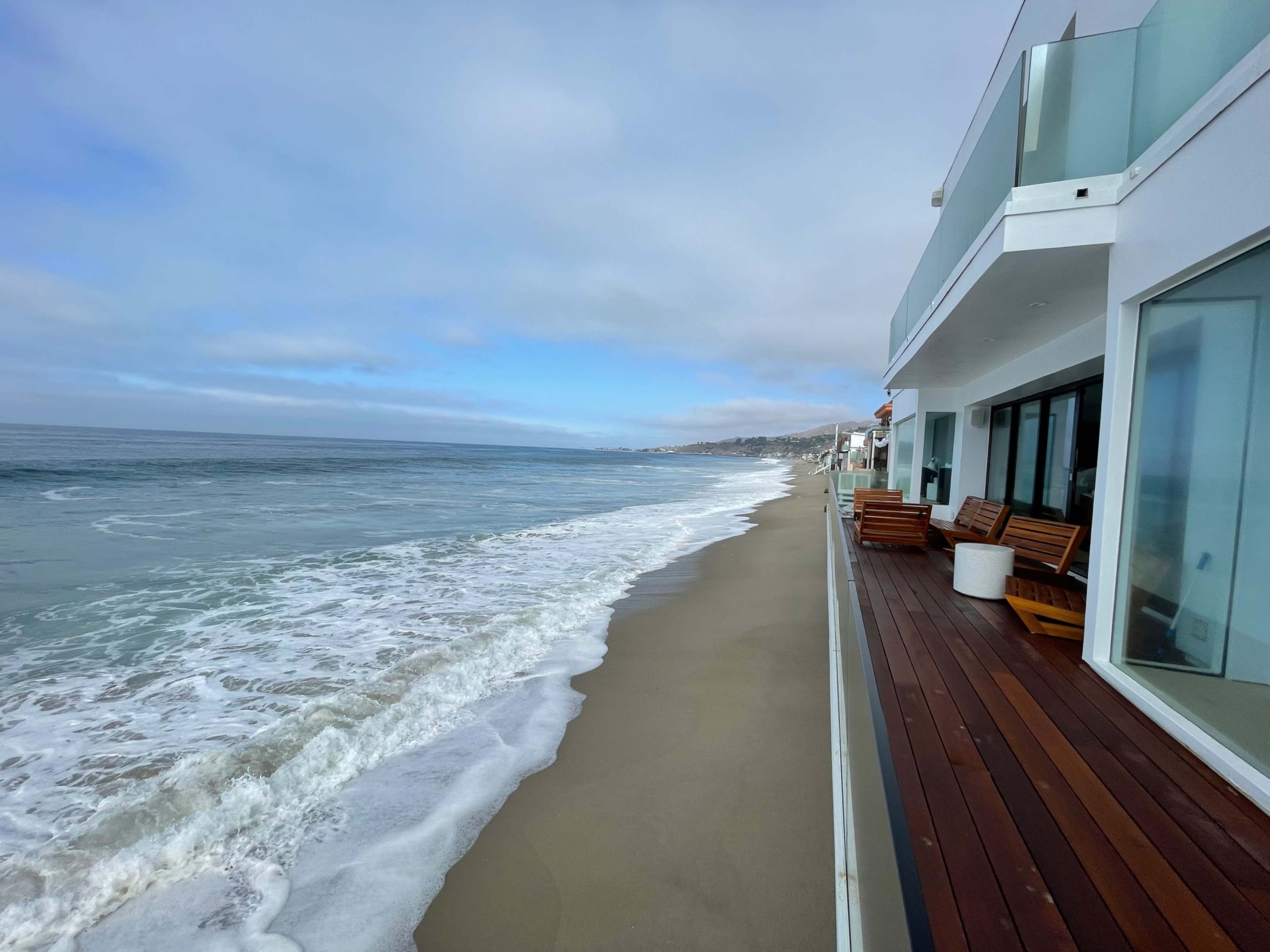 A modern beachfront property overlooks a wide sandy shoreline and gentle waves under a cloudy sky.