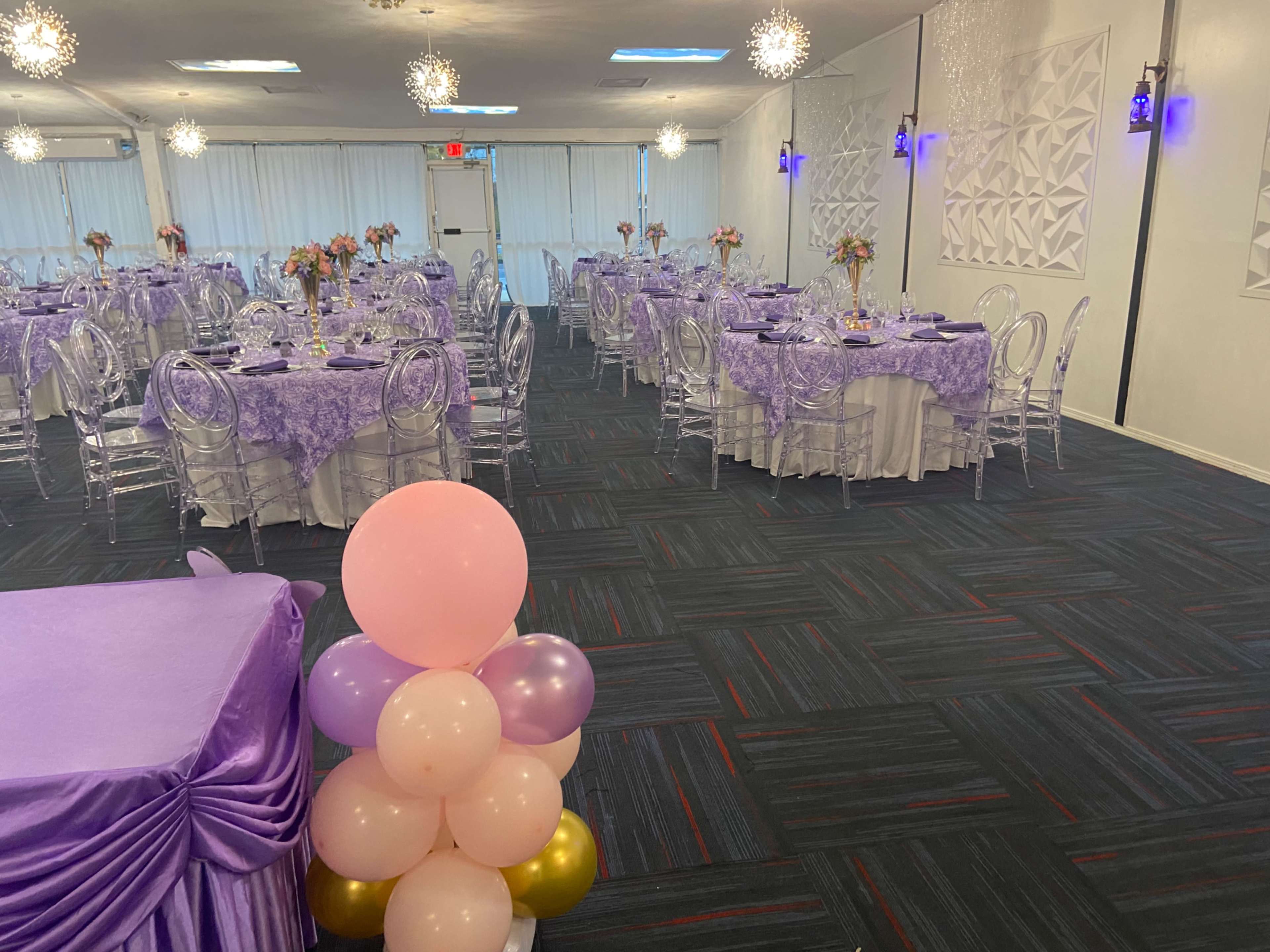 The image shows a banquet hall set up for an event, featuring tables with purple tablecloths and clear acrylic chairs, decorated with floral centerpieces.