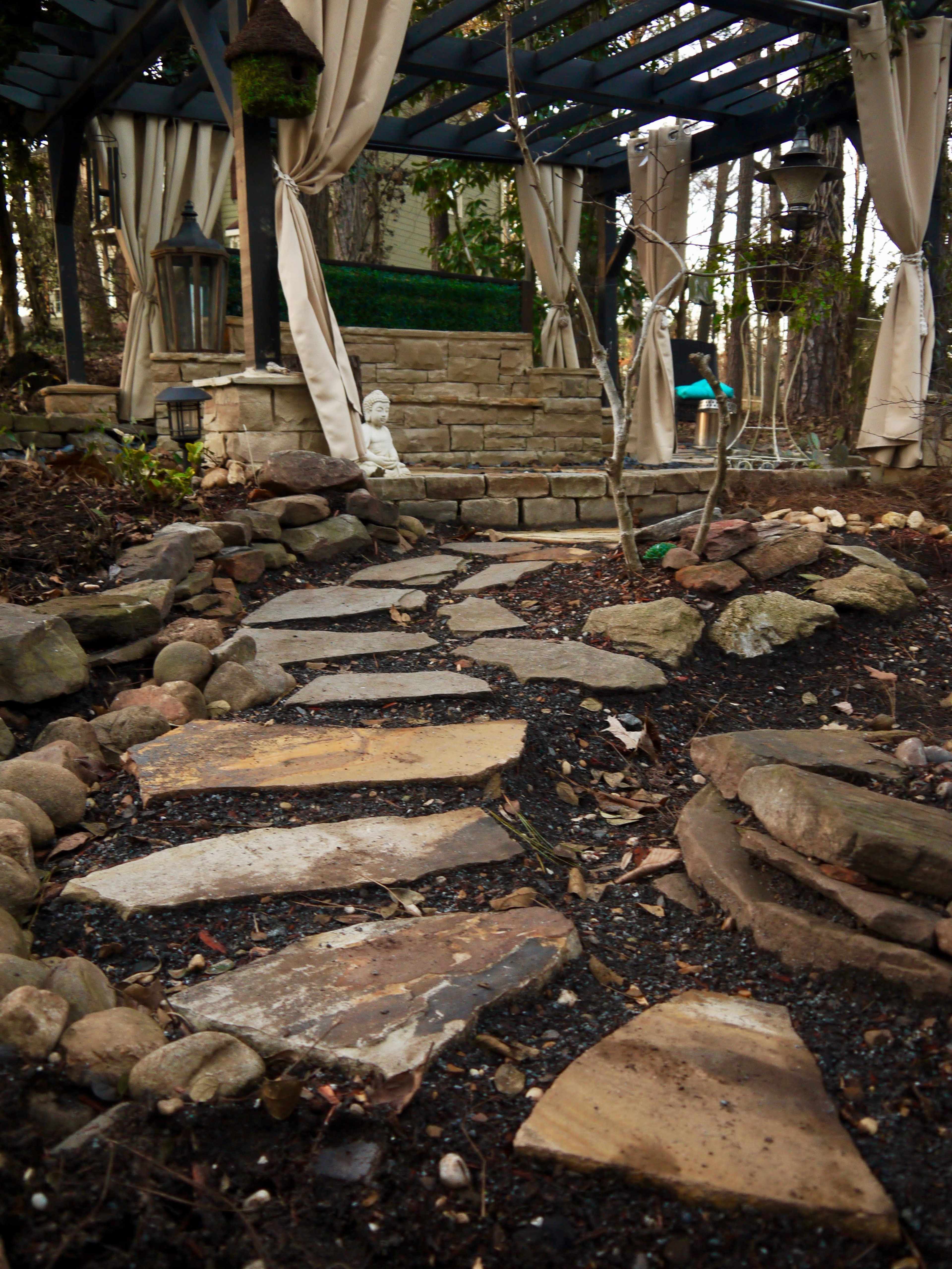 A stone pathway leads through a landscaped garden with a stone seating area under a wooden pergola.