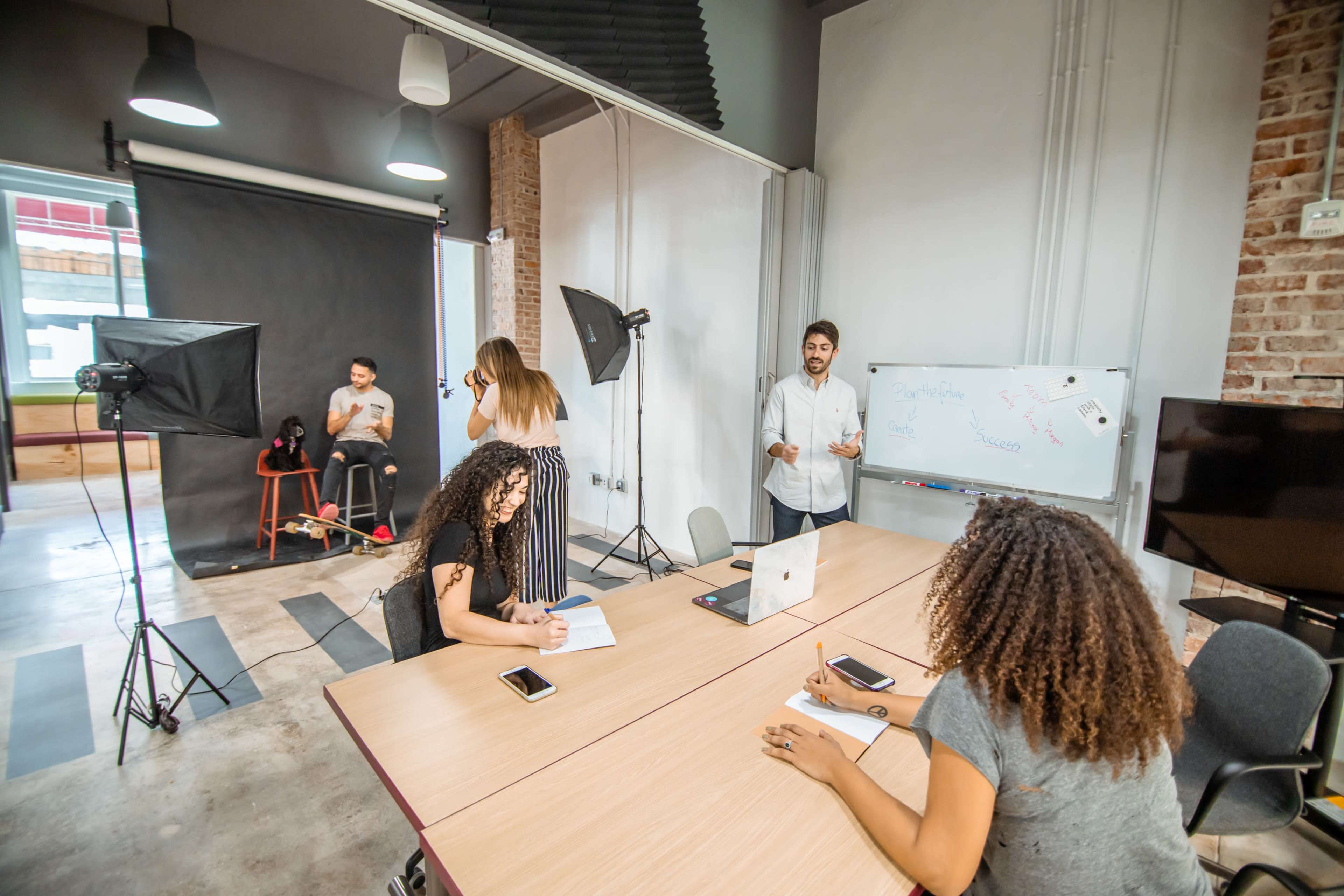 A group of people engage in a collaborative work session in a well-lit studio featuring a large table, a whiteboard, and photography equipment.