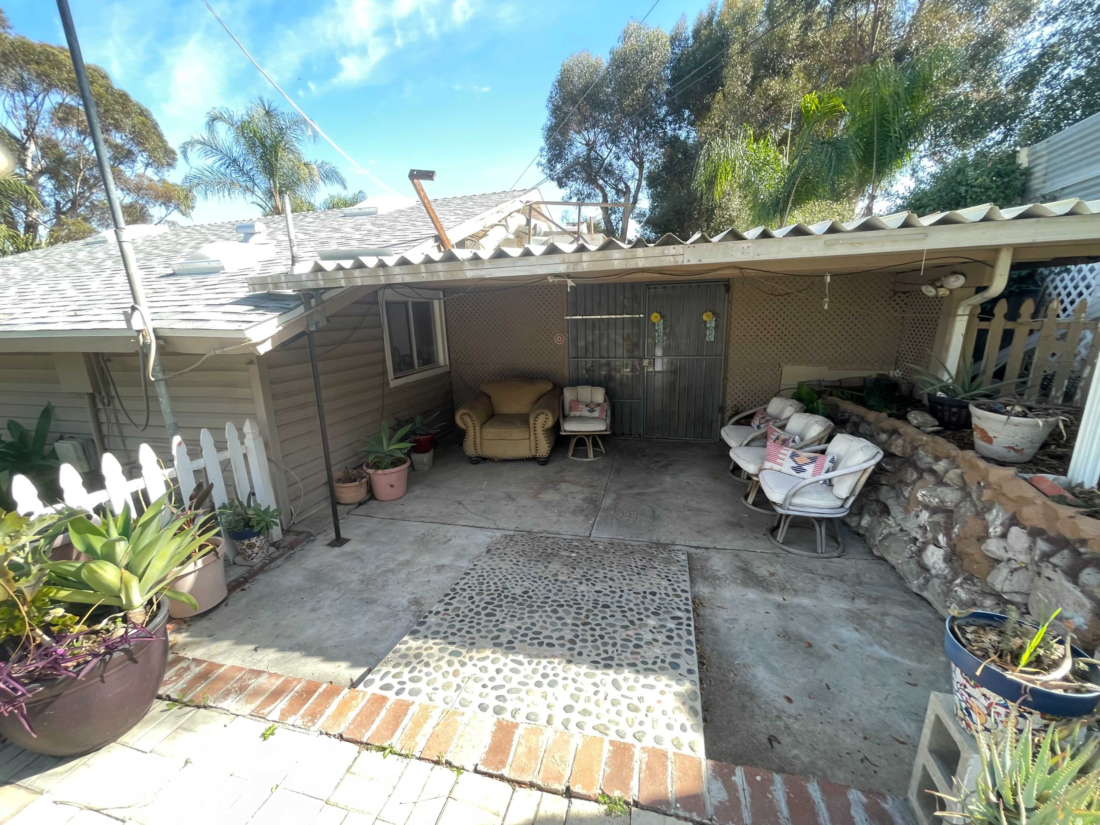 The image features a small outdoor patio area with a concrete floor, potted plants, and seating arrangements, set against a house that has a sloped roof and a white picket fence.