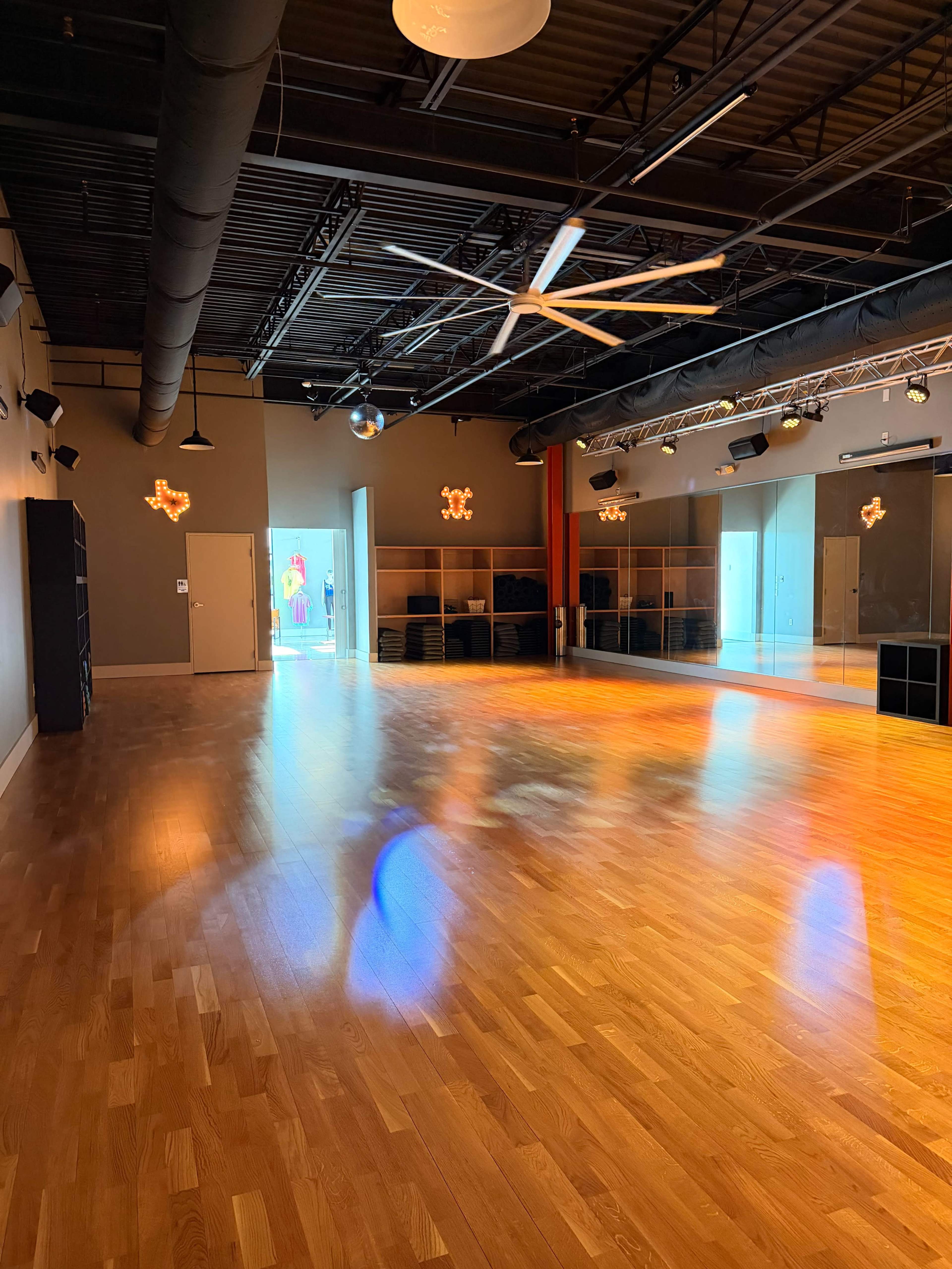 The image shows an empty dance studio with wooden flooring, large mirrors, and bright overhead lighting.