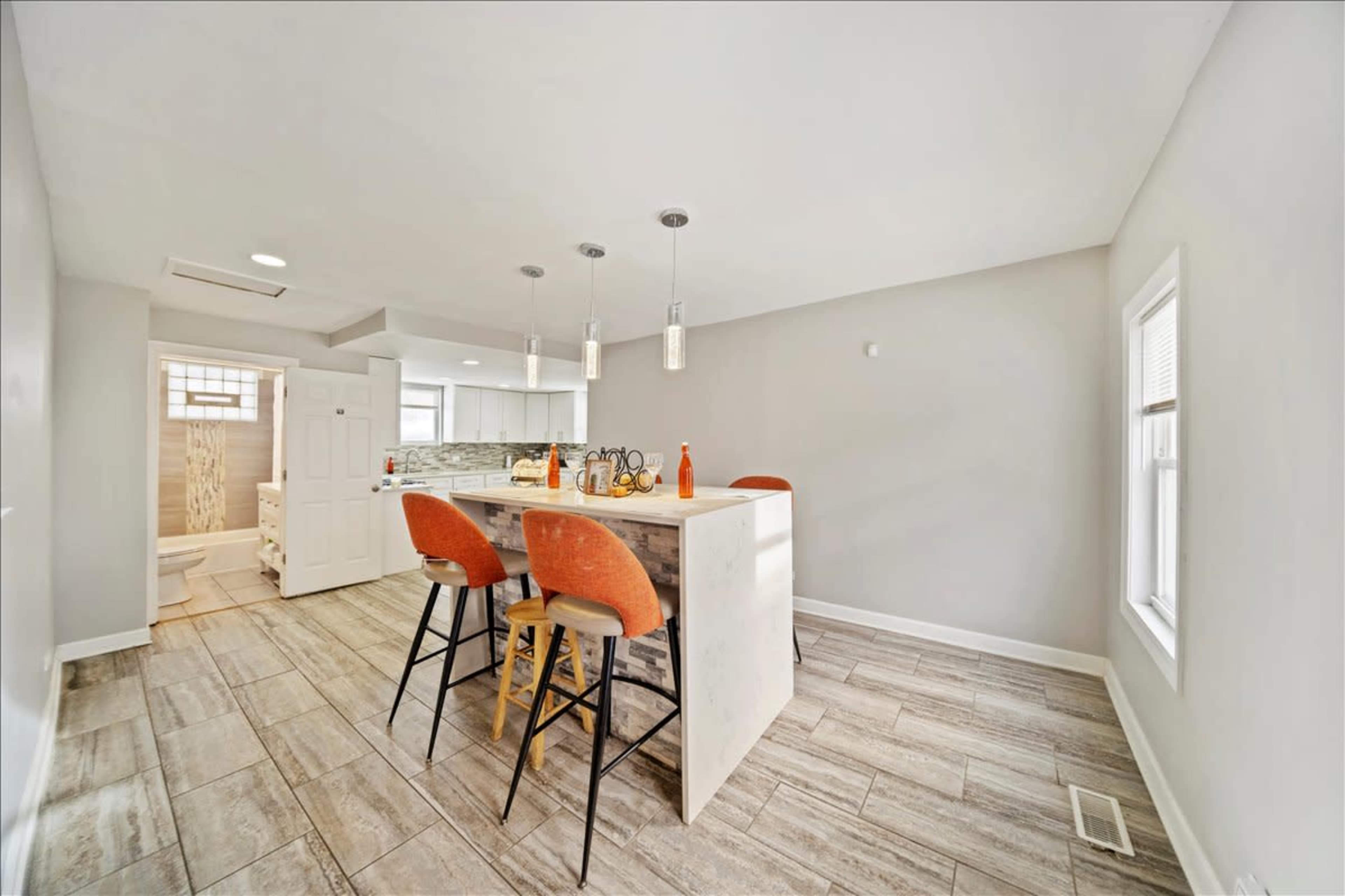 The image shows a modern kitchen area featuring a central island with four orange bar stools and a doorway leading to a bathroom.