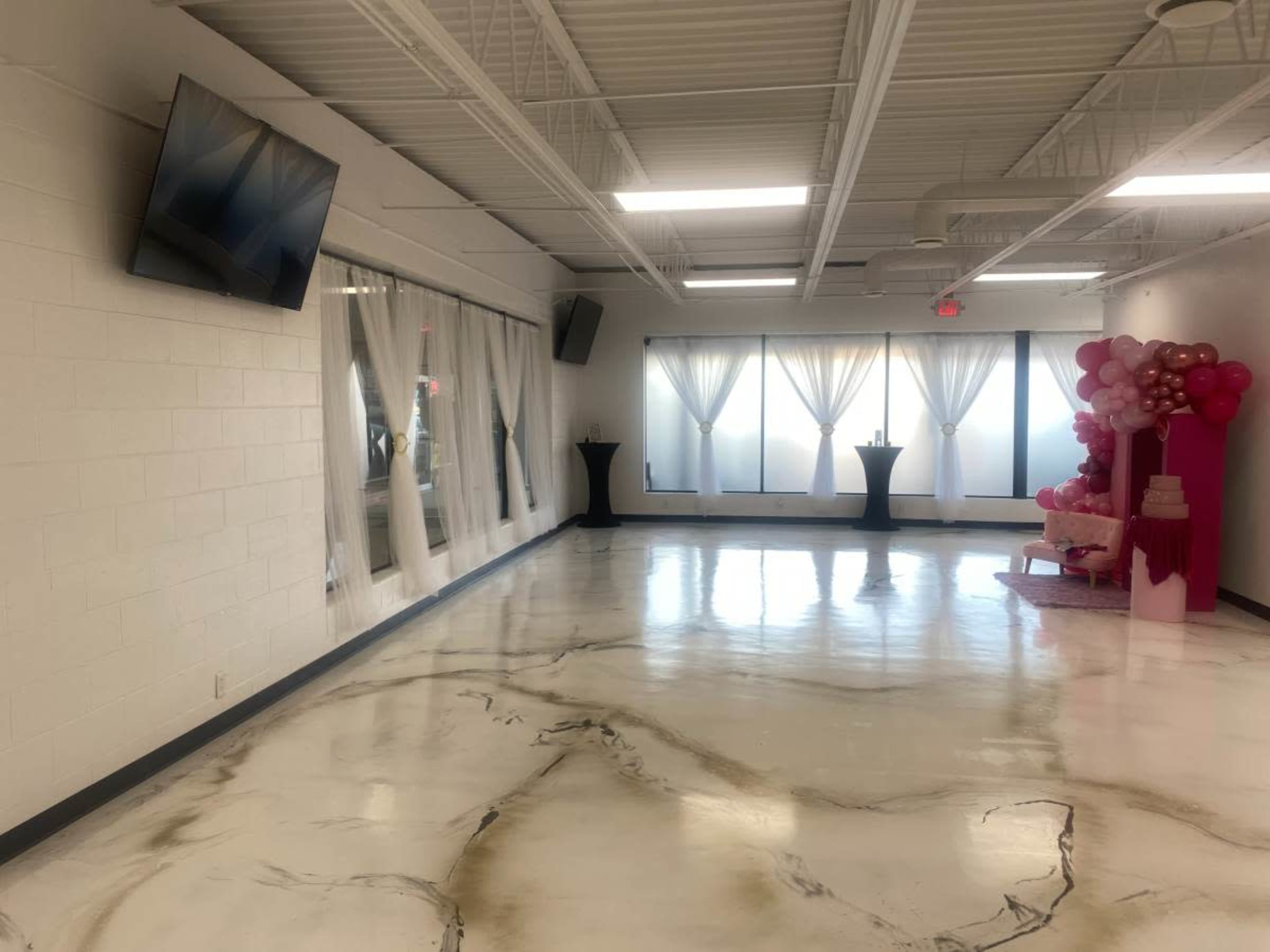 The image shows a spacious room with polished floors, large windows covered by sheer curtains, and minimal decorations, including two black tables and a pink balloon arrangement in one corner.