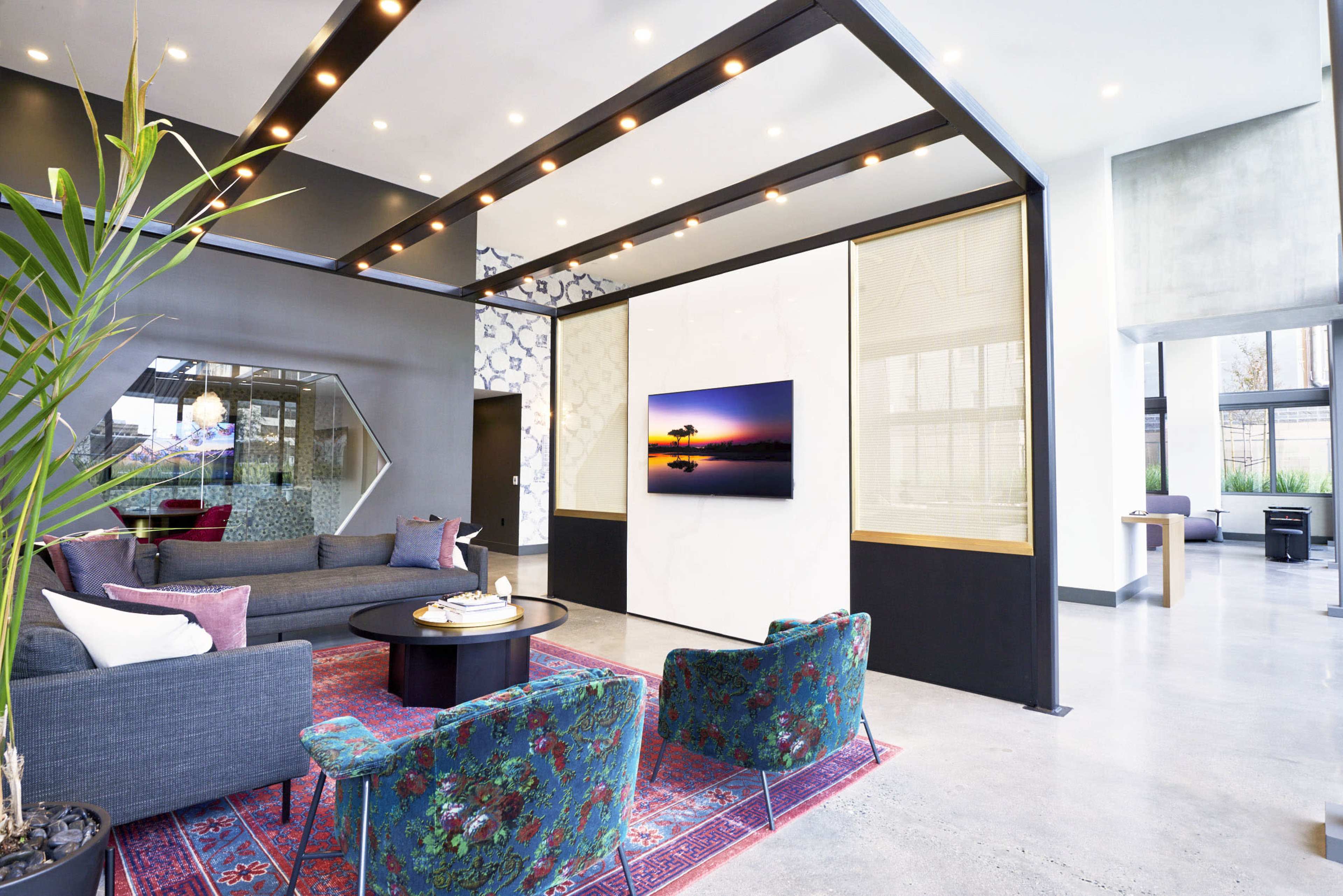 The image shows a modern lounge area featuring a large TV mounted on a white wall, a dark sofa, two patterned chairs, and decorative plants.