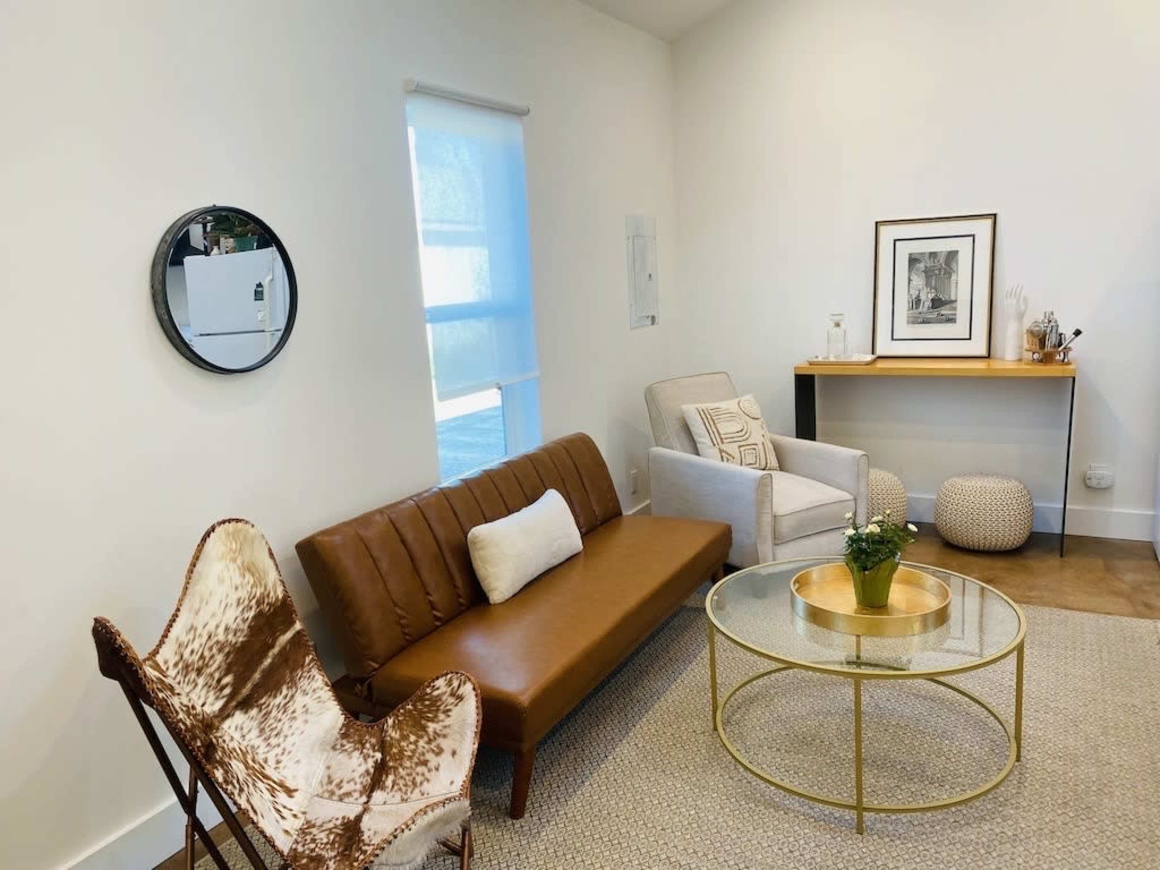 A modern living space features a brown sofa, a beige armchair, a round glass coffee table, and a small writing desk against a light-colored wall.