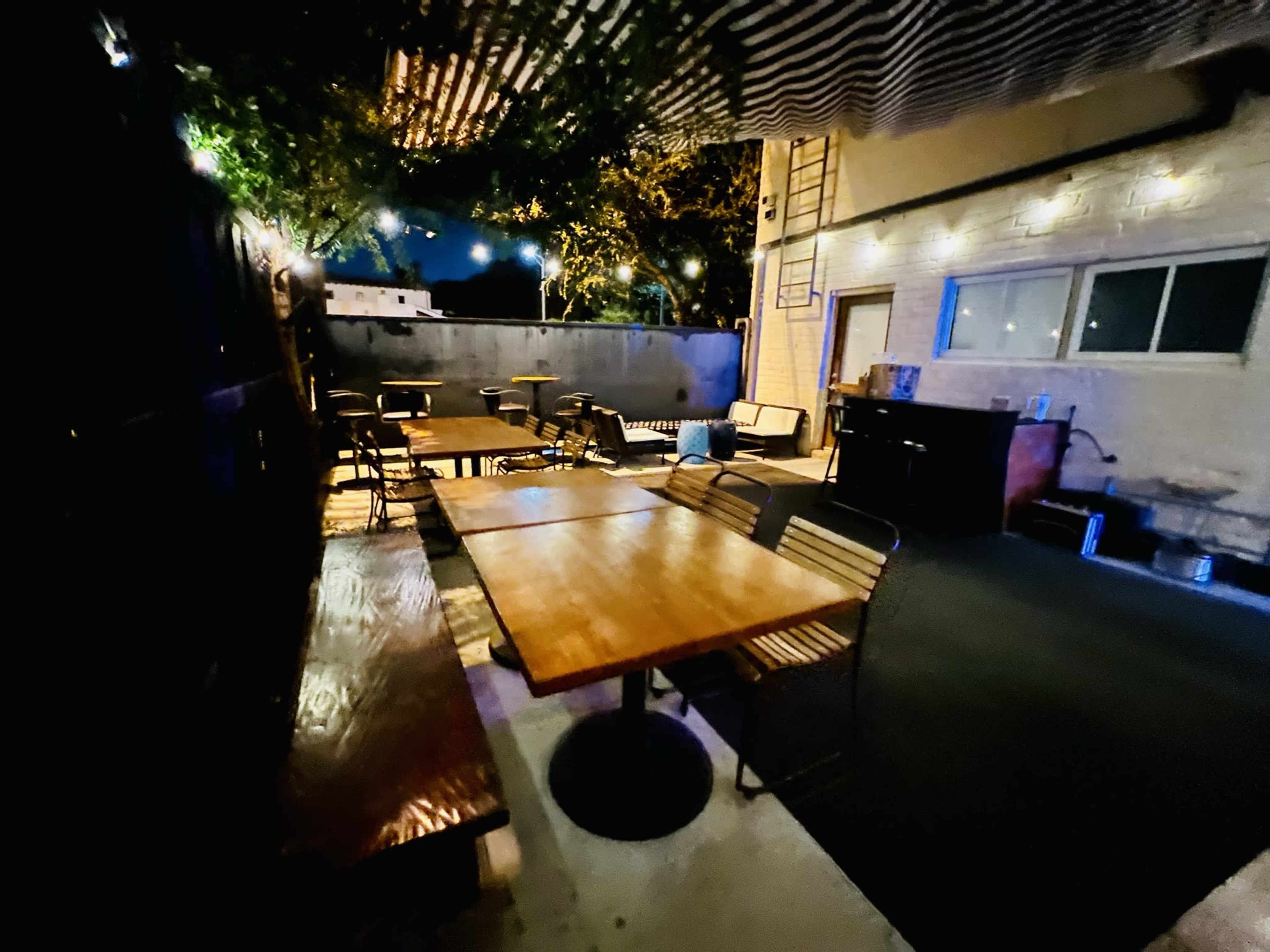 An outdoor seating area with several wooden tables and chairs under string lights and a partially shaded overhead structure.