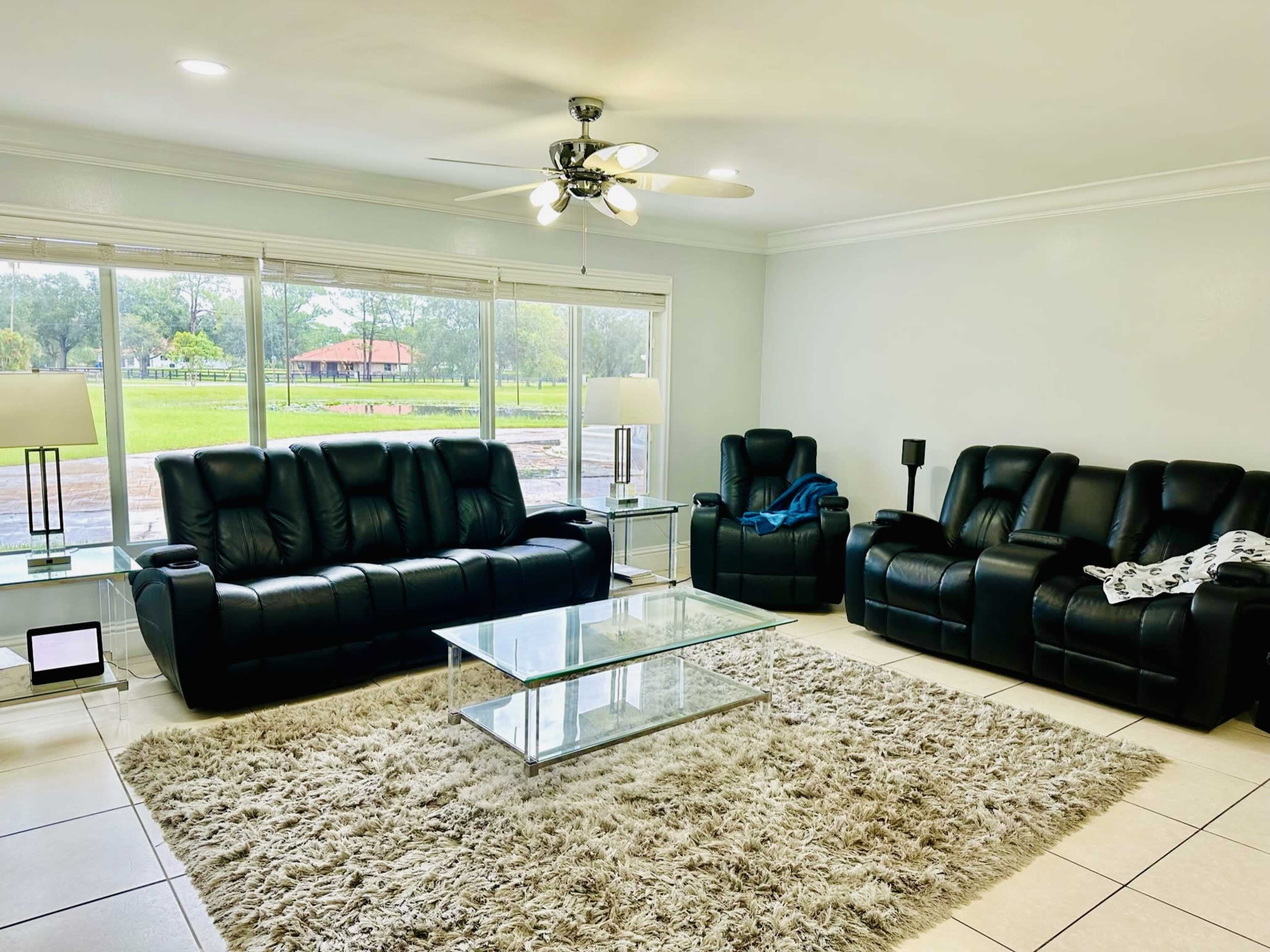 A living room with black leather sofas, a glass coffee table, and large windows overlooking a green outdoor area.