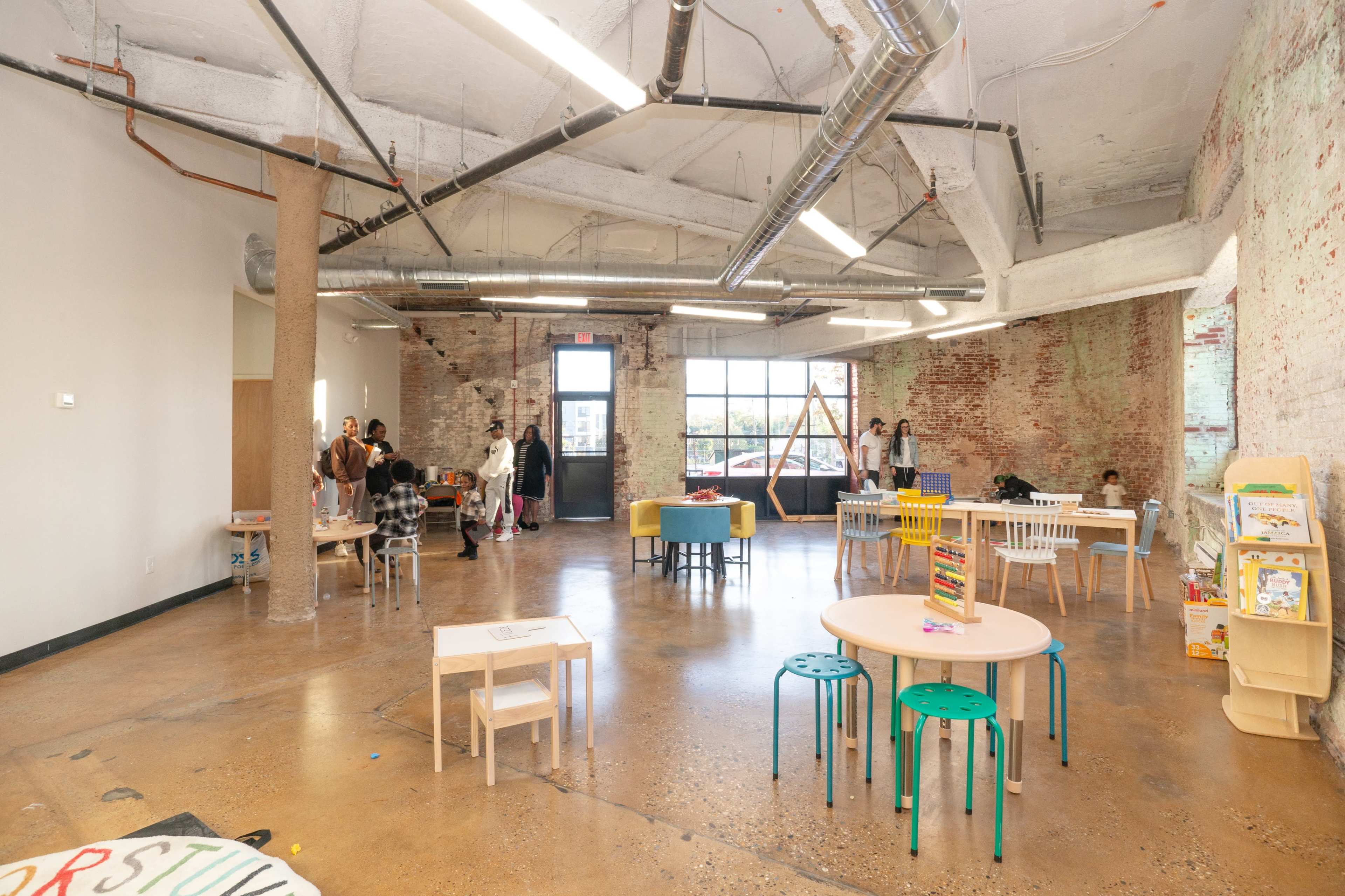 A spacious, modern classroom features several tables and chairs, with children and adults engaging in activities near large windows and exposed brick walls.