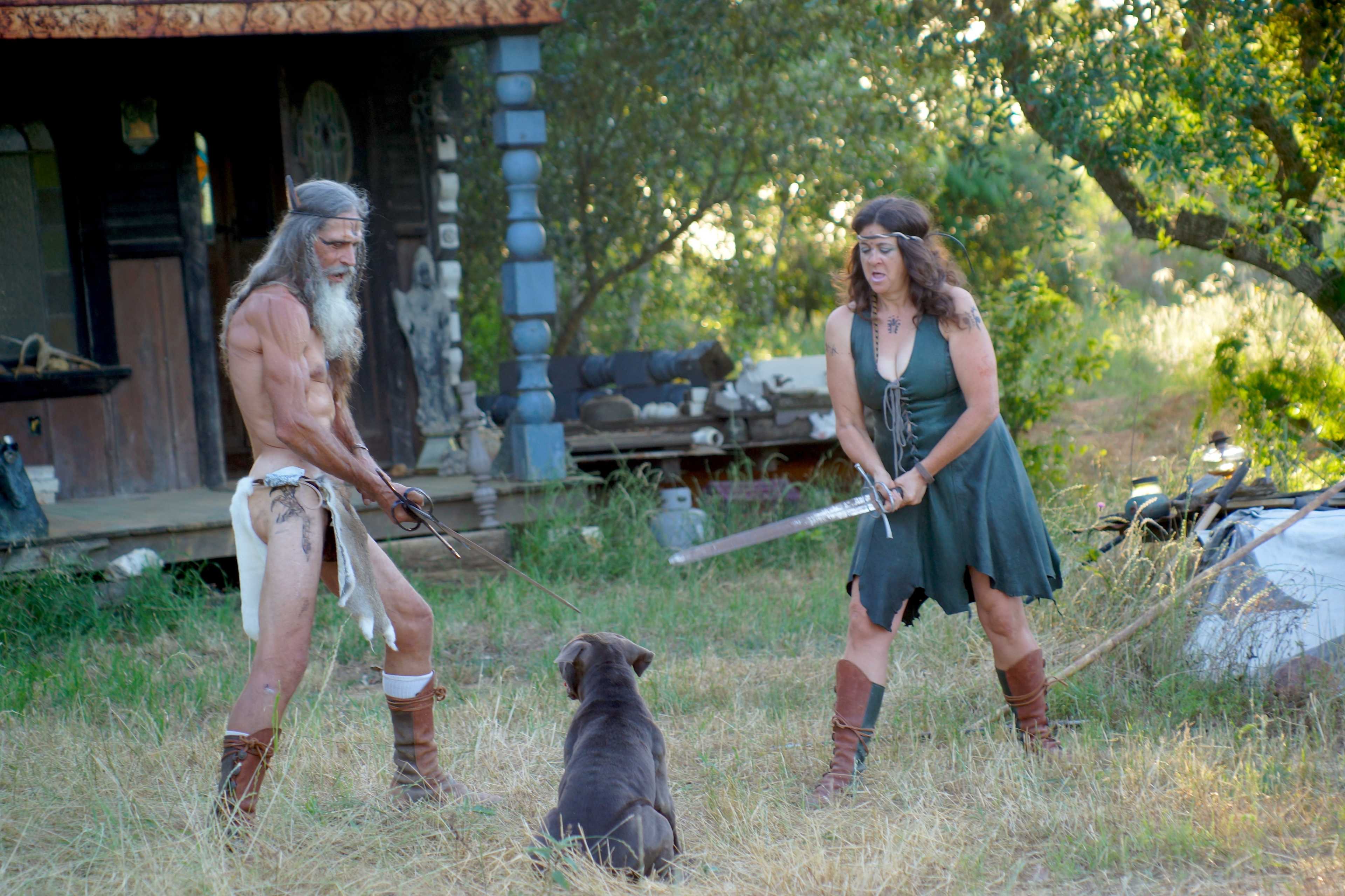 A man and a woman, both dressed in primitive attire, stand facing each other with swords while a dog sits between them in a grassy area near a rustic cabin.