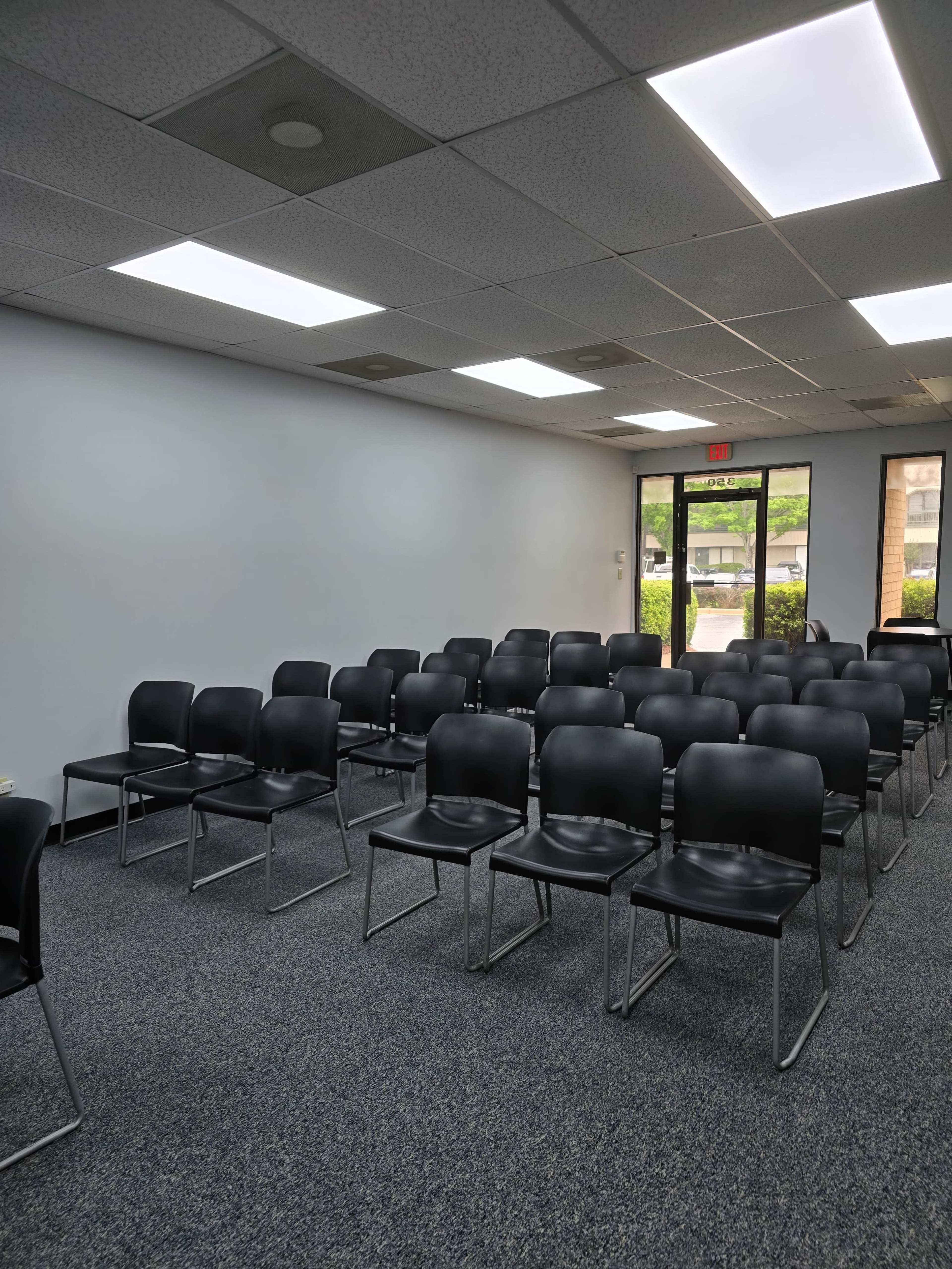 The image shows a room with rows of empty black chairs arranged facing a door and windows, with a gray carpet and white walls.