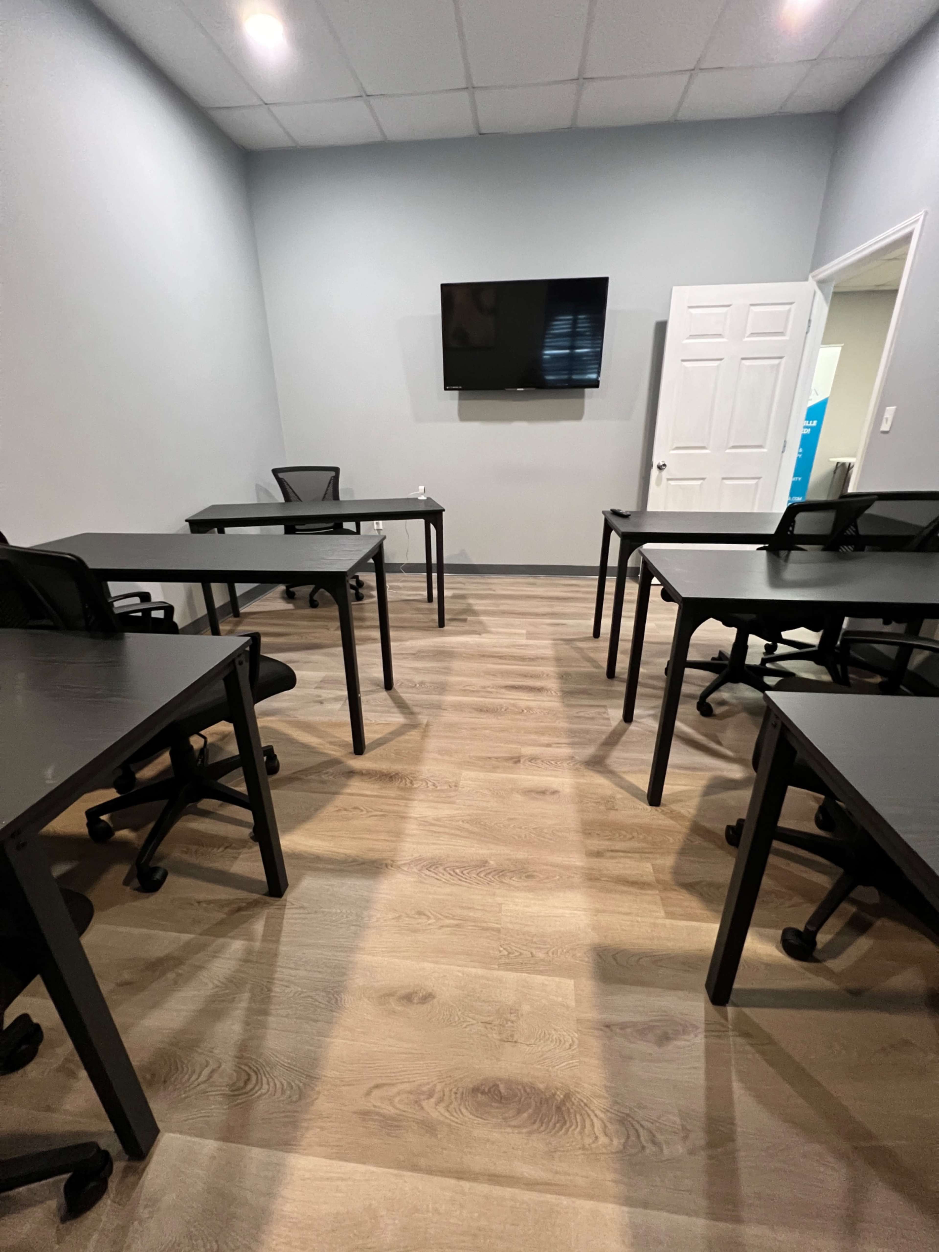 A meeting room with several black desks and chairs arranged around a central open space, featuring a wall-mounted television and a white door.