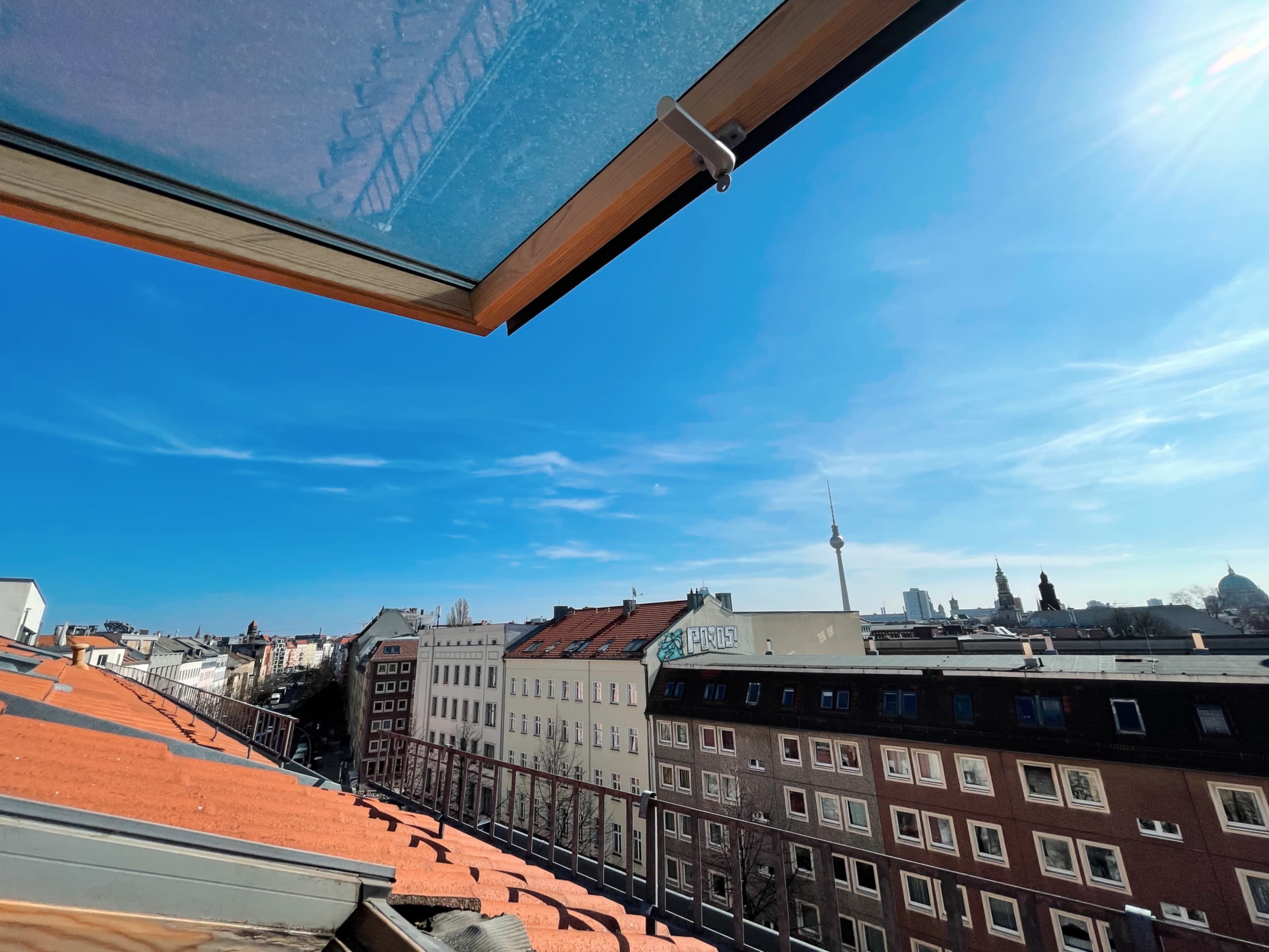 The image shows a view from a rooftop featuring red-tiled buildings and a clear blue sky, with a tower in the distance.